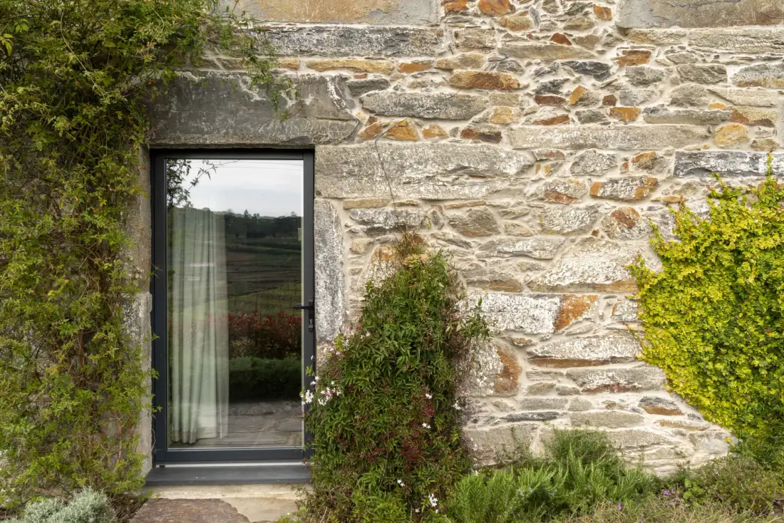Una pared exterior de piedra con una puerta de cristal rodeada de vegetación.