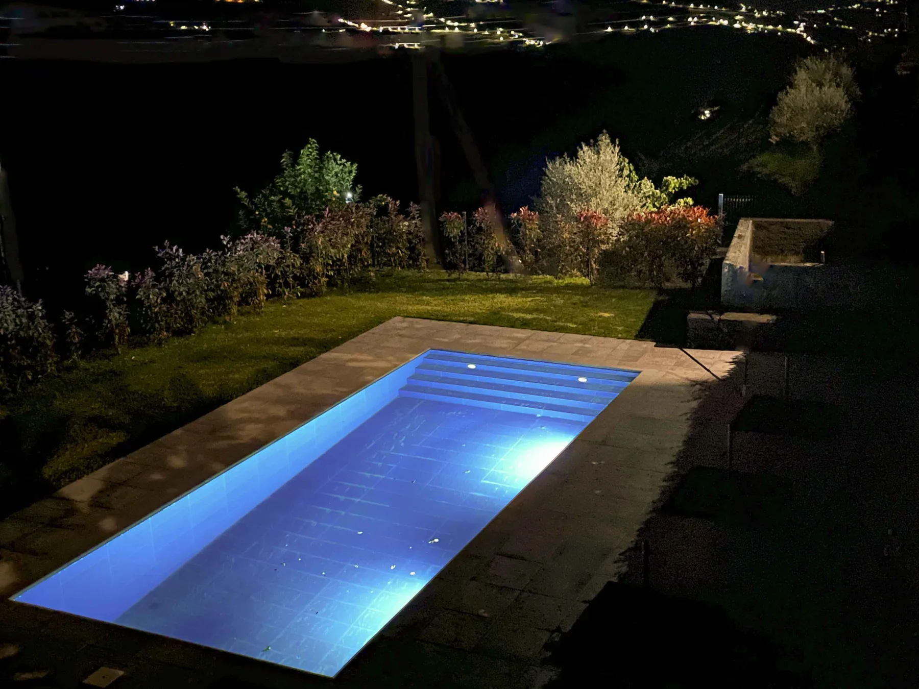 Una piscina iluminada de azul junto a un jardín ajardinado por la noche.