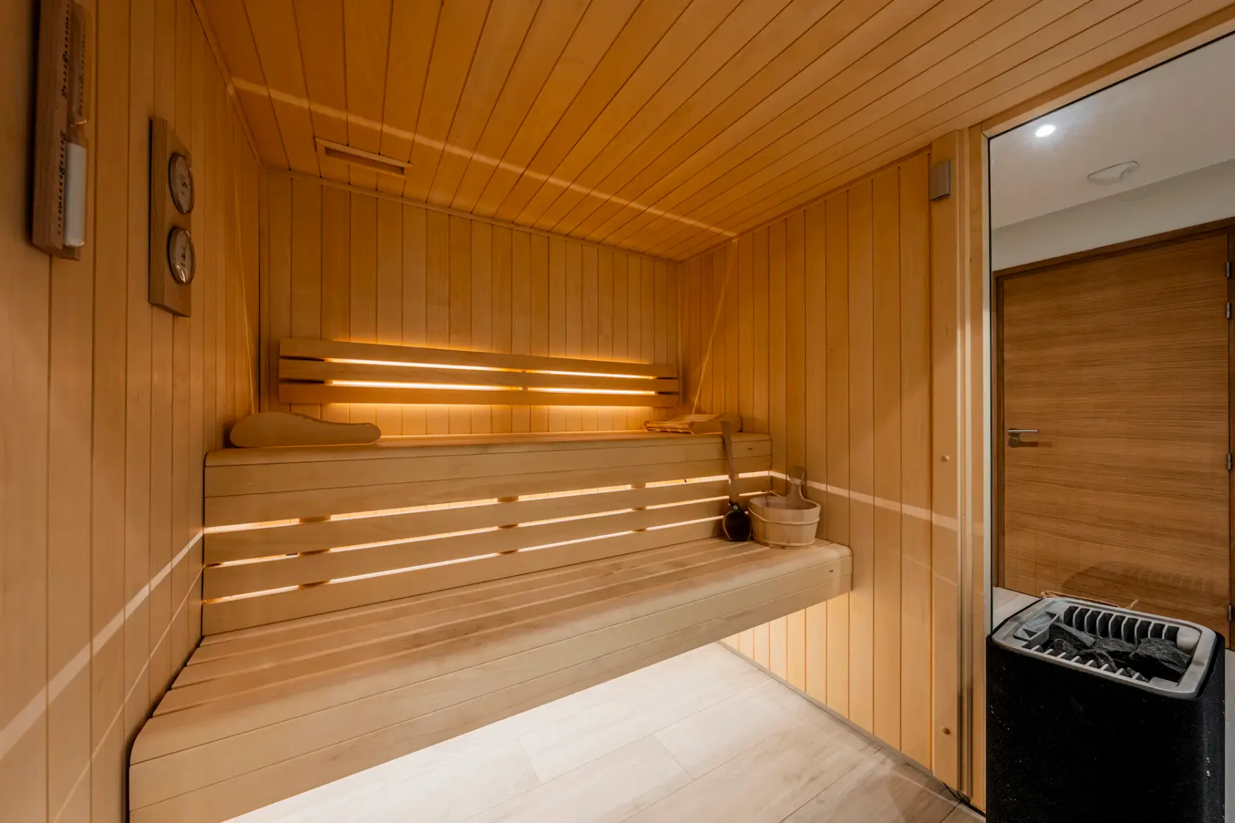 A wooden sauna room with tiered benches, a bucket and ladle, and a heater. Warm lighting creates a cozy atmosphere.
