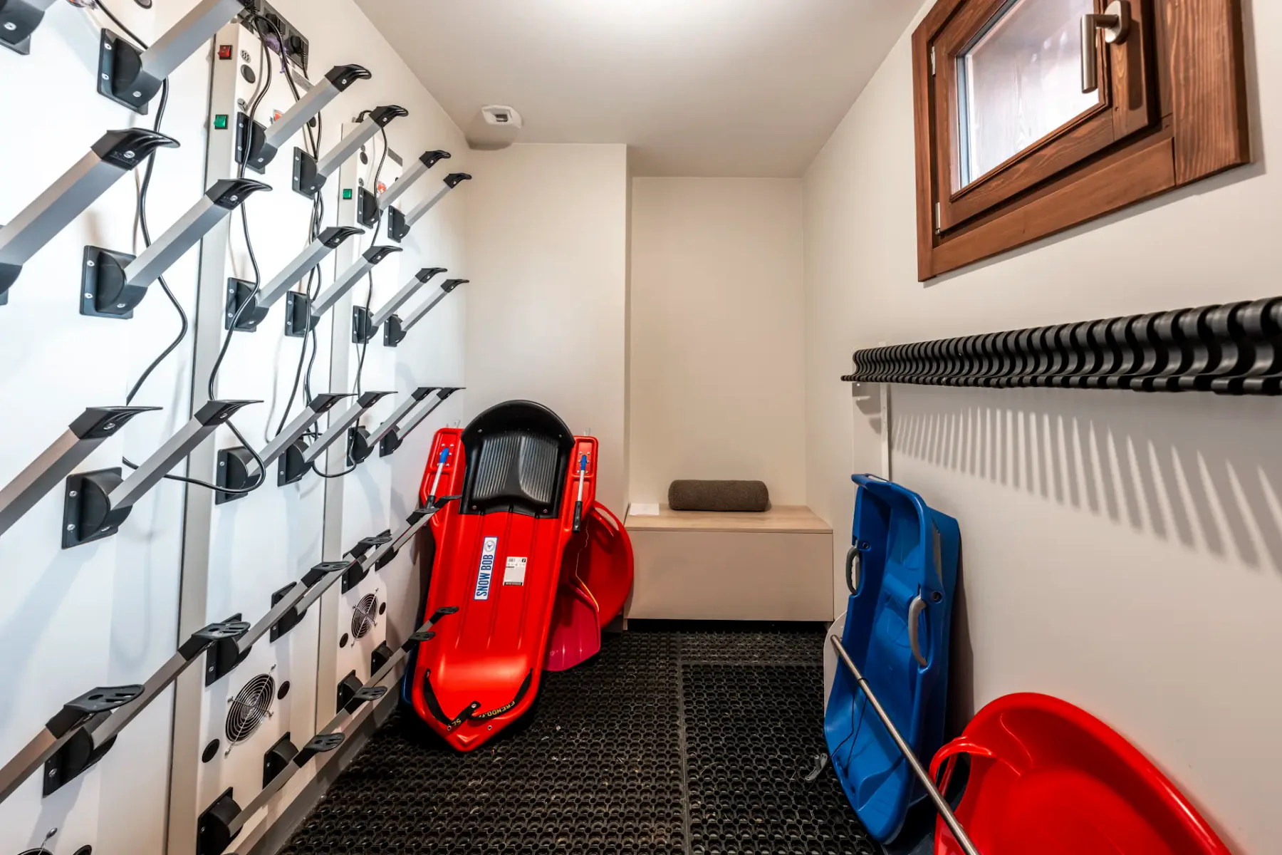 Room with wall-mounted boot dryers, red and blue sleds, a bench, and a small window.