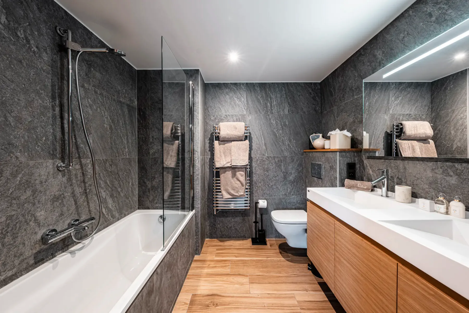 Modern bathroom with dark stone walls, wood flooring, a bathtub, large mirror, and a wooden vanity with a white countertop.