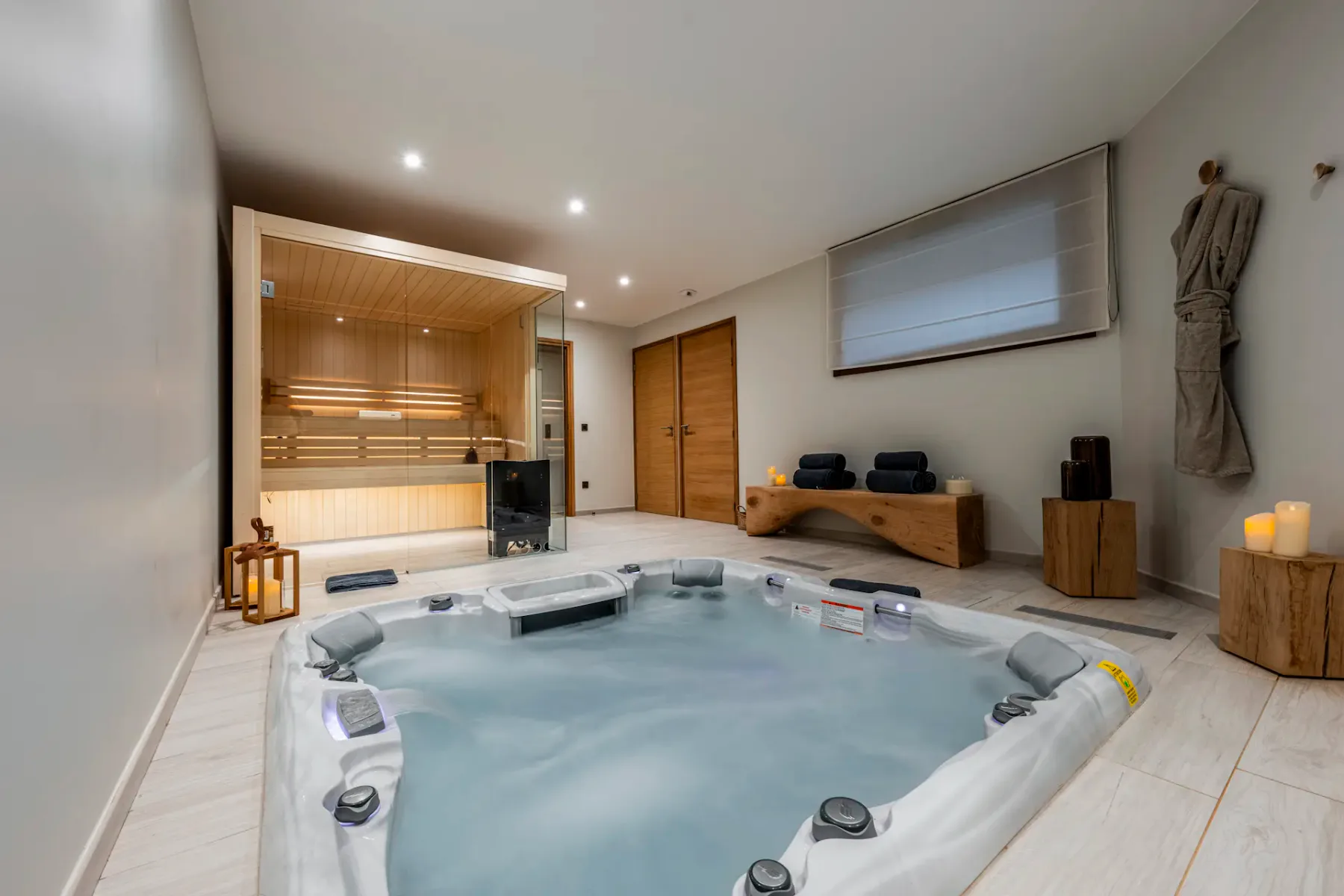 A modern spa room with a jacuzzi, wooden sauna, candles, and folded towels on a bench, creating a serene and relaxing atmosphere.