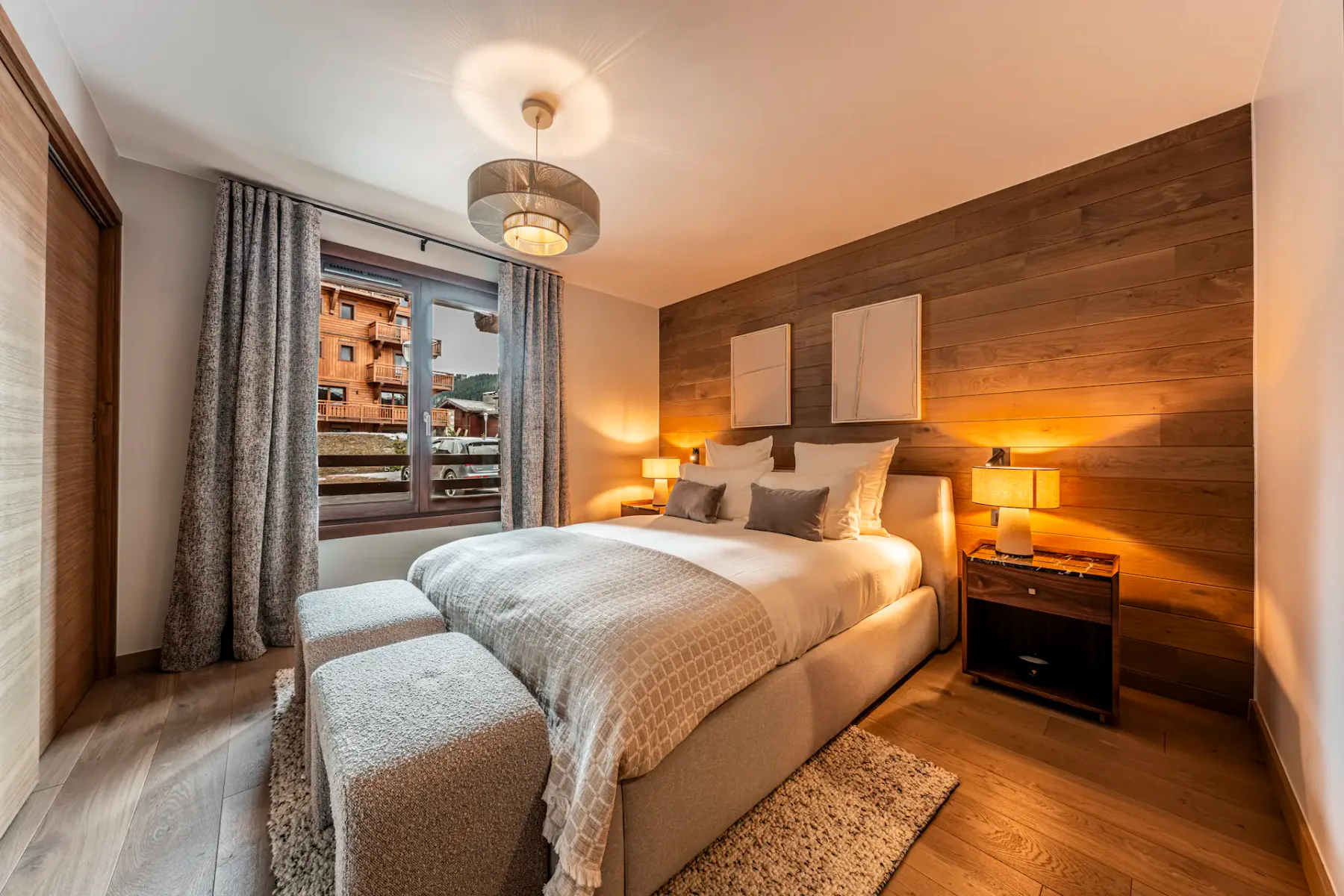 Cozy bedroom with a double bed, wooden accent wall, soft lighting, and a window view of buildings. Decor includes a rug and cushioned ottomans.