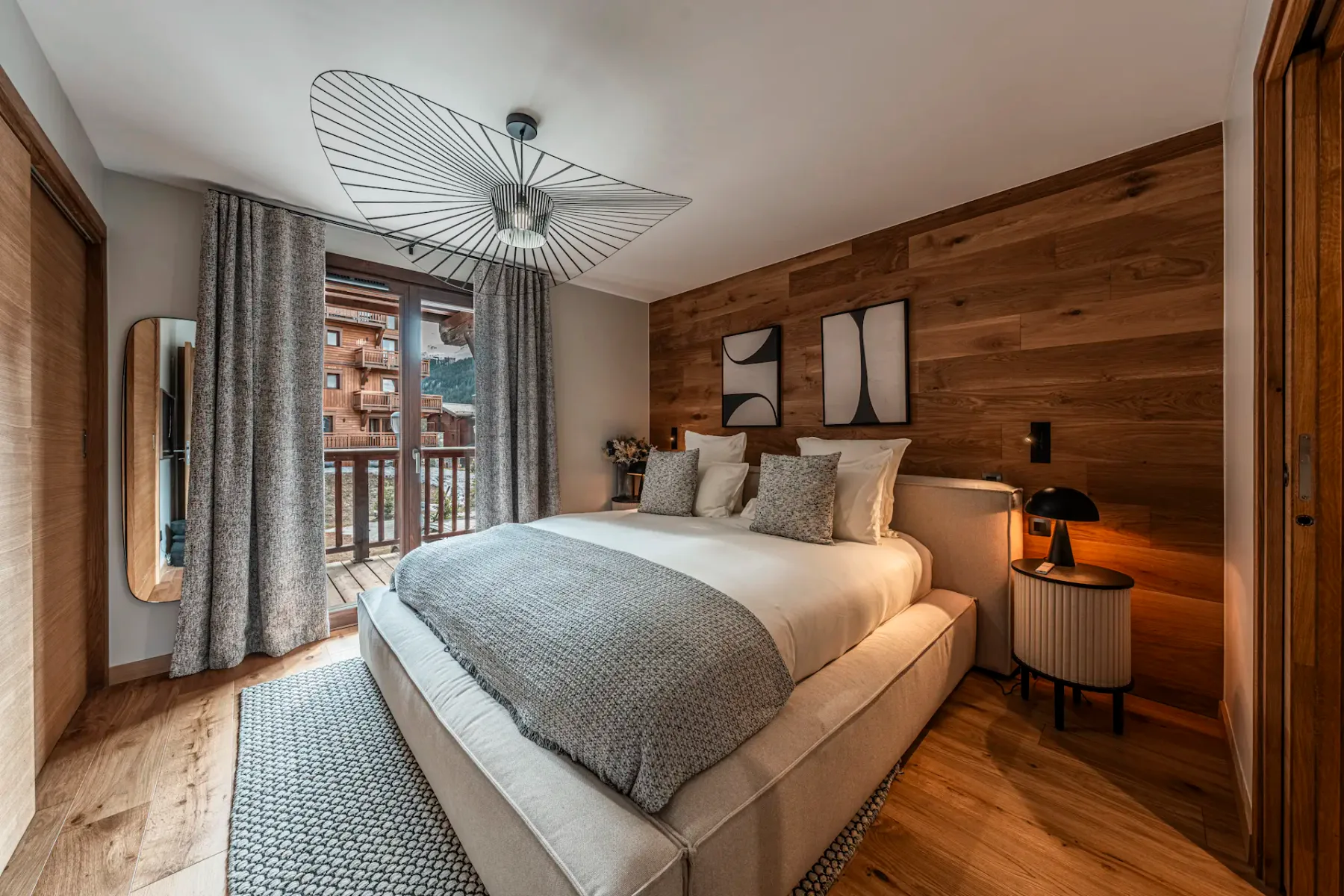Cozy bedroom with a large bed, wooden accent wall, modern lighting, and a balcony view. Neutral tones and stylish decor create a warm atmosphere.