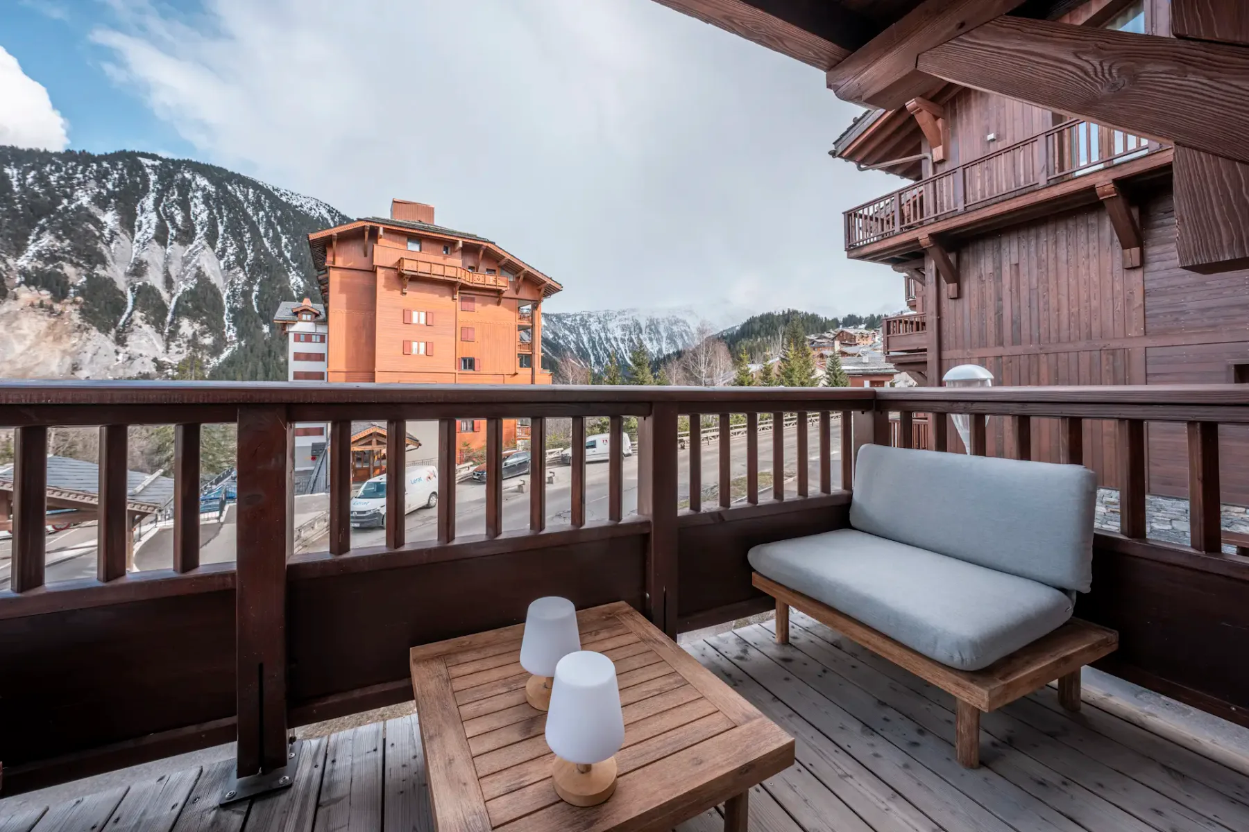 A wooden balcony with a cushioned bench, table, and two lamps overlooks a mountain view and nearby buildings.