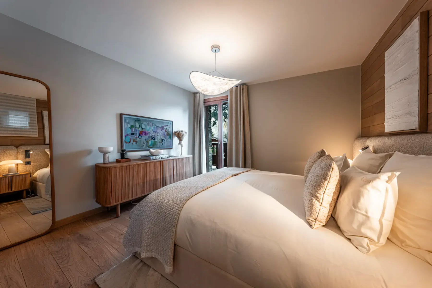 Cozy bedroom with a large bed, wooden dresser, wall mirror, and modern lighting. A window with curtains lets in natural light.