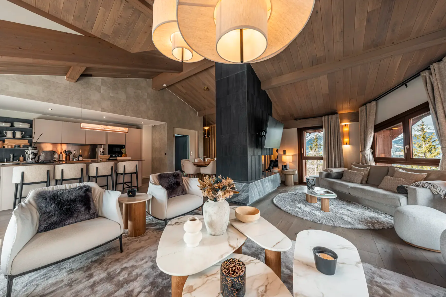 Spacious modern living room with wooden ceiling, cozy seating, marble tables, and large windows. Open kitchen with bar stools in the background.