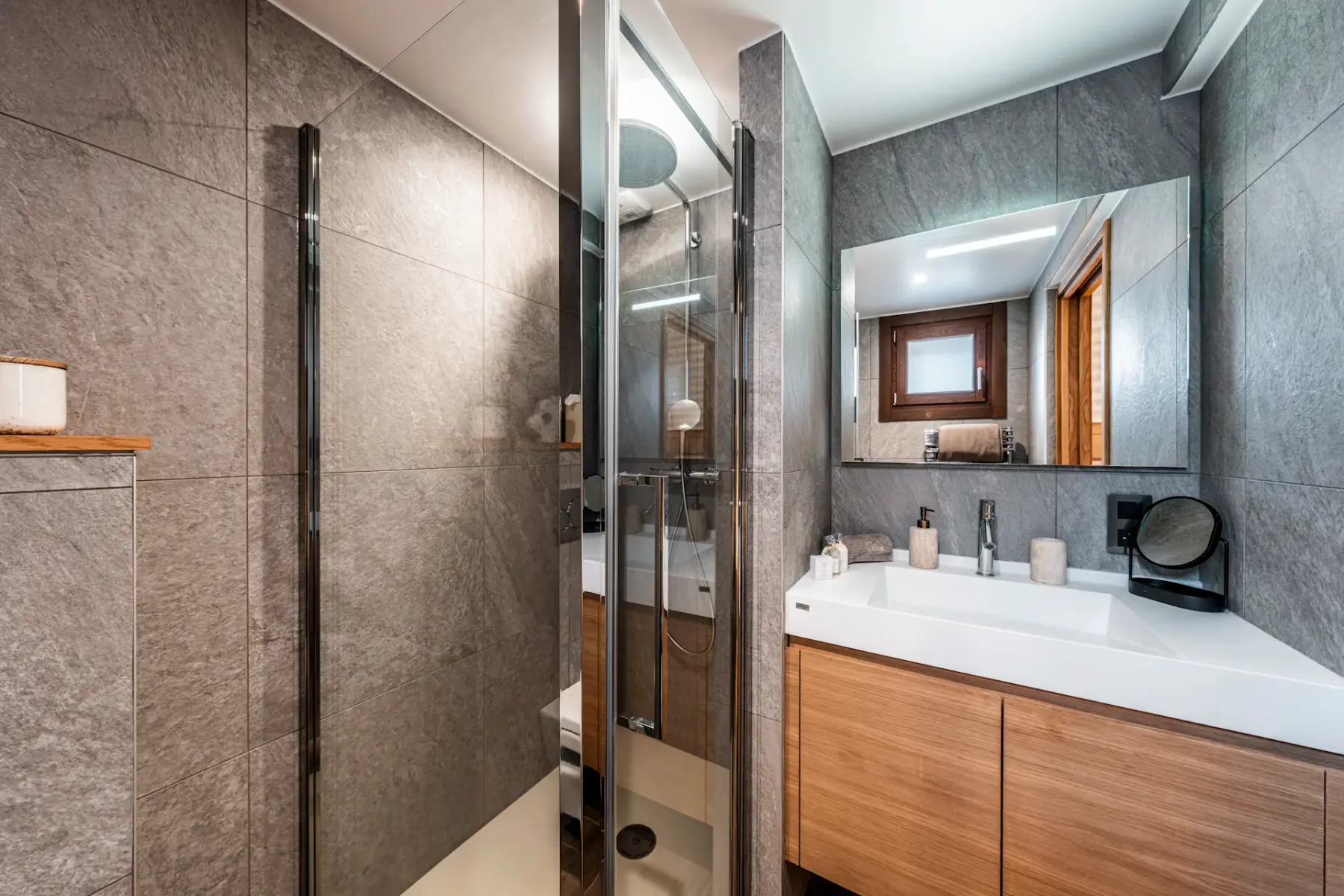 Modern bathroom with gray tiled walls, glass shower, wooden vanity with a white sink, mirror, and small window. Minimalist decor.