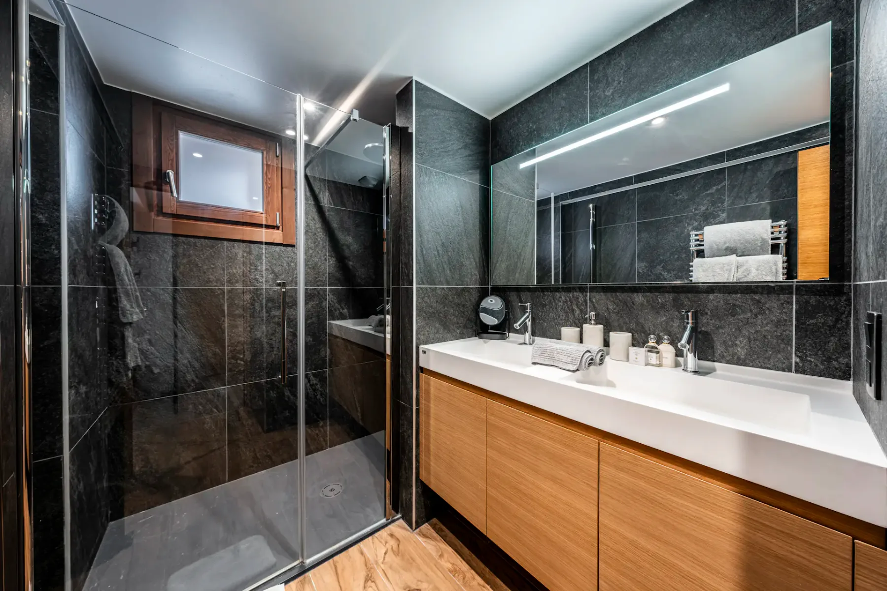 Modern bathroom with dark tiles, a glass shower enclosure, wooden vanity with a white countertop, large mirror, and a small window.