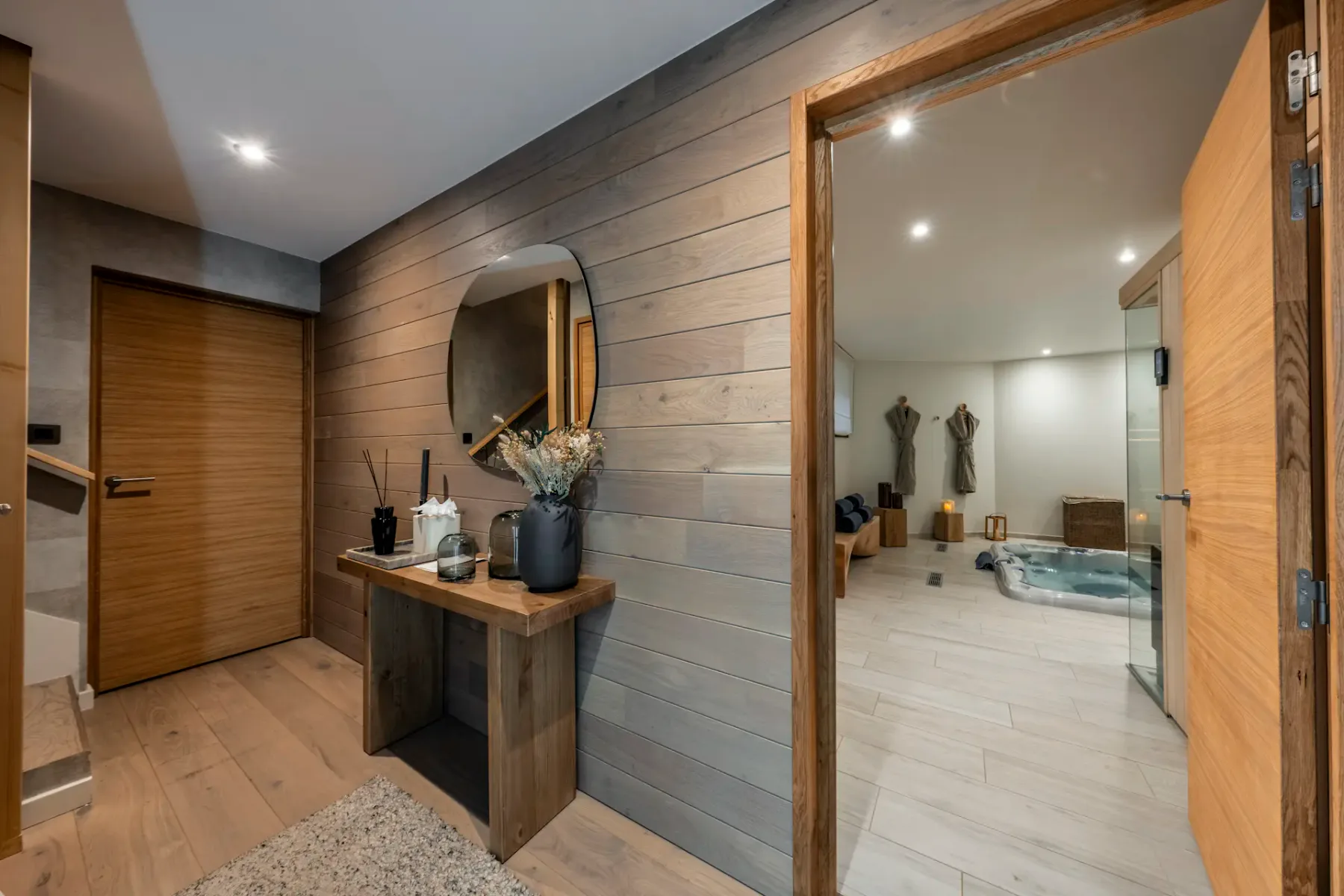 Modern interior with wood accents, featuring a hallway with a round mirror and decorative items, leading to a cozy room with seating and soft lighting.