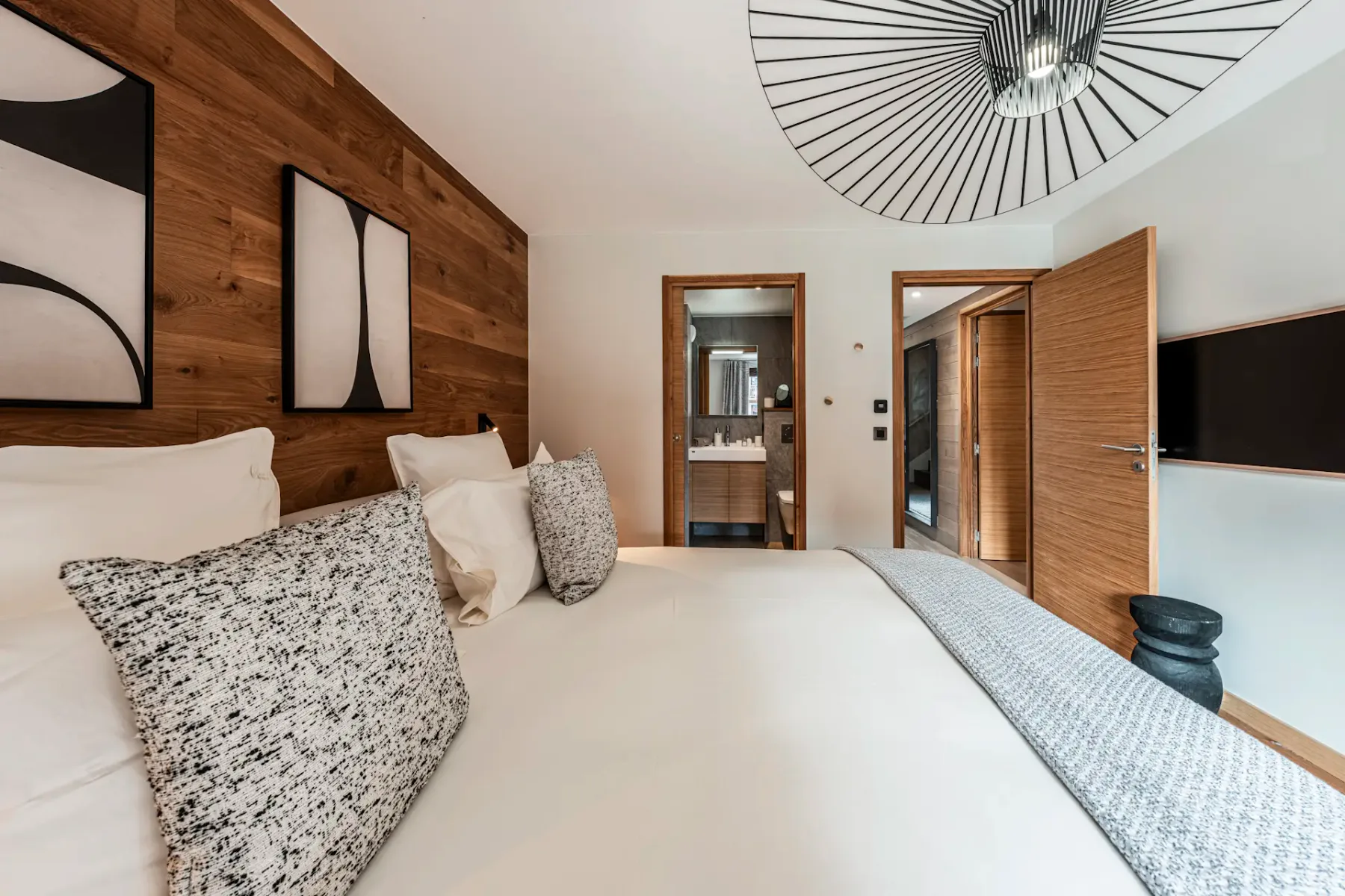 Modern bedroom with a large bed, patterned pillows, wooden accent wall, abstract art, and open doors leading to a bathroom and hallway.
