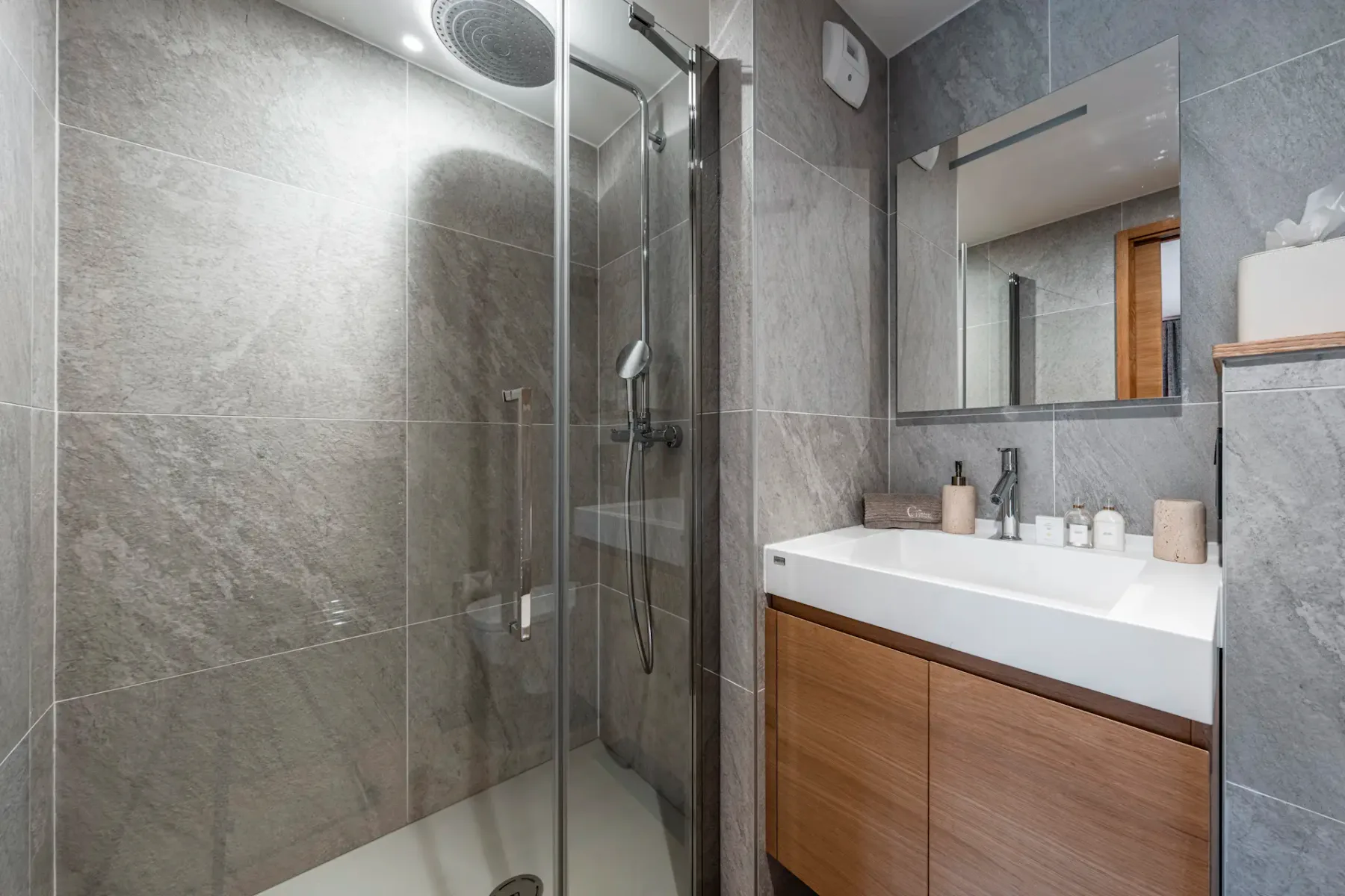 Modern bathroom with a glass-enclosed shower, gray tiled walls, a wooden vanity with a white sink, and a large mirror.