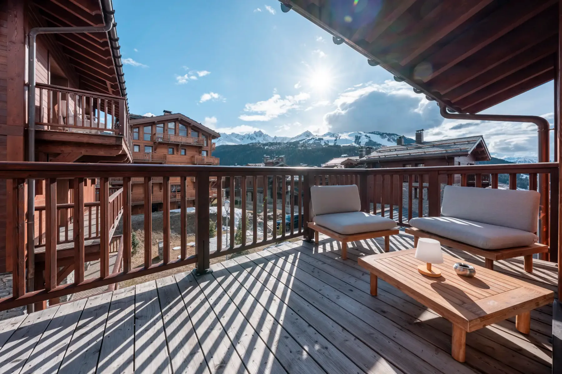 Sunny mountain view from a wooden balcony with cozy seating, surrounded by rustic wooden buildings and snow-capped peaks in the distance.