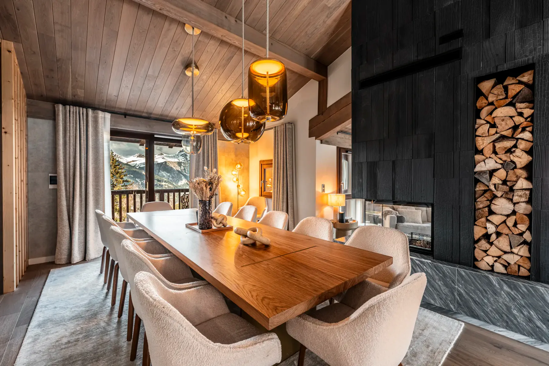 Modern dining room with a long wooden table, plush chairs, pendant lights, and a fireplace with stacked logs. Large windows offer mountain views.