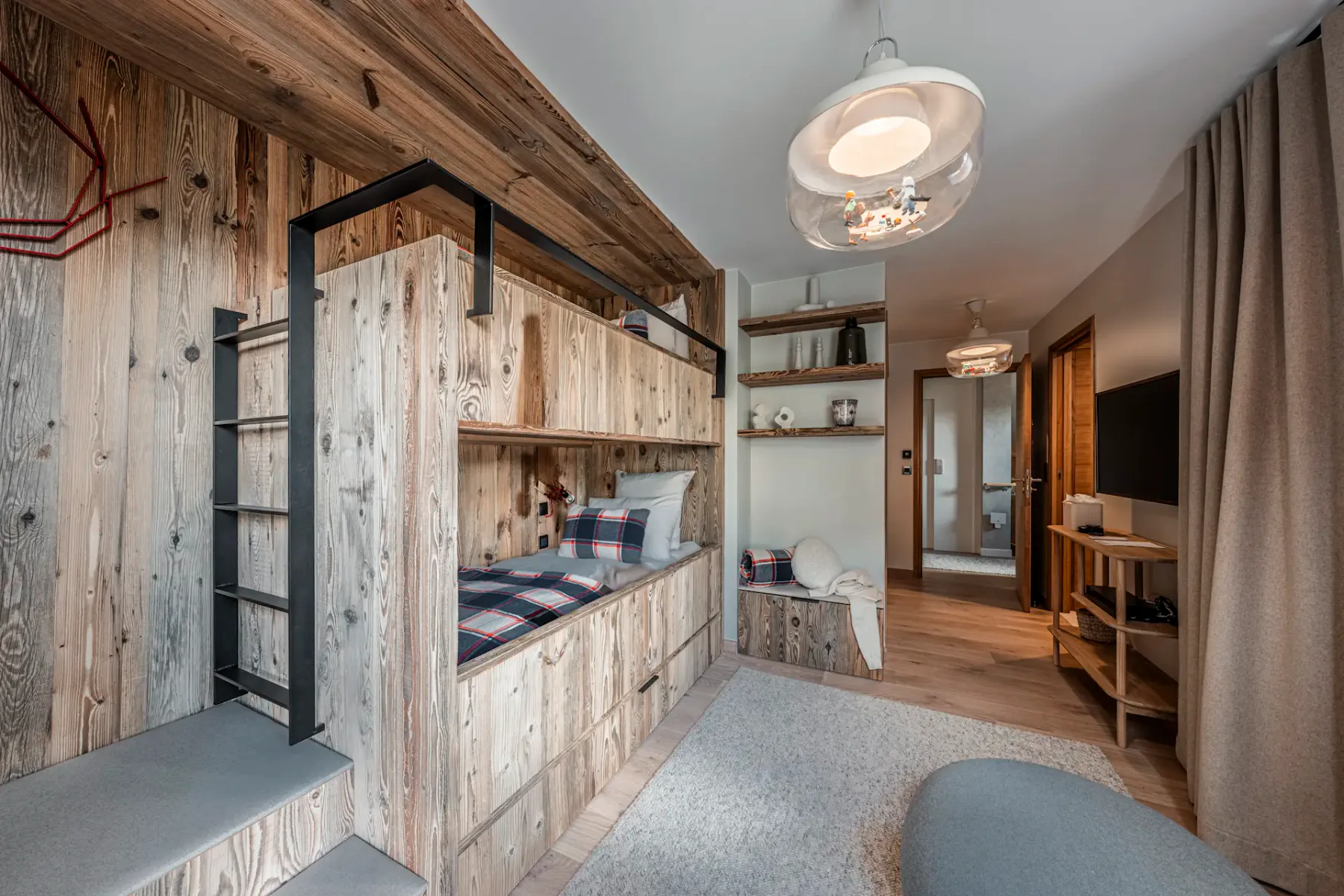 Cozy room with wooden bunk bed, plaid pillows, and modern lighting. Shelves and a TV are visible, with neutral decor and soft lighting.