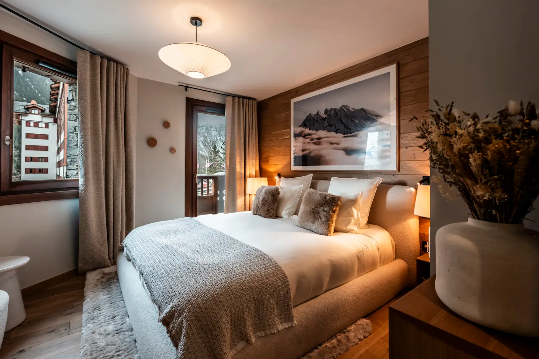 Cozy bedroom with a large bed, plush pillows, soft lighting, mountain artwork, and a window view of snowy scenery. Warm, inviting atmosphere.