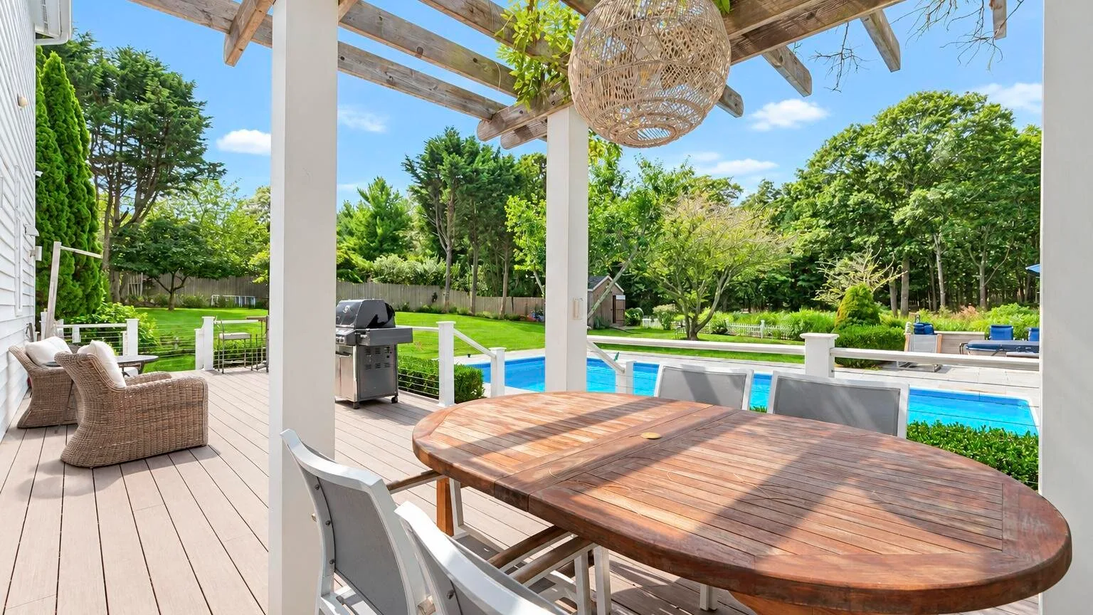 Una mesa de comedor de madera está bajo una pérgola junto a una piscina.