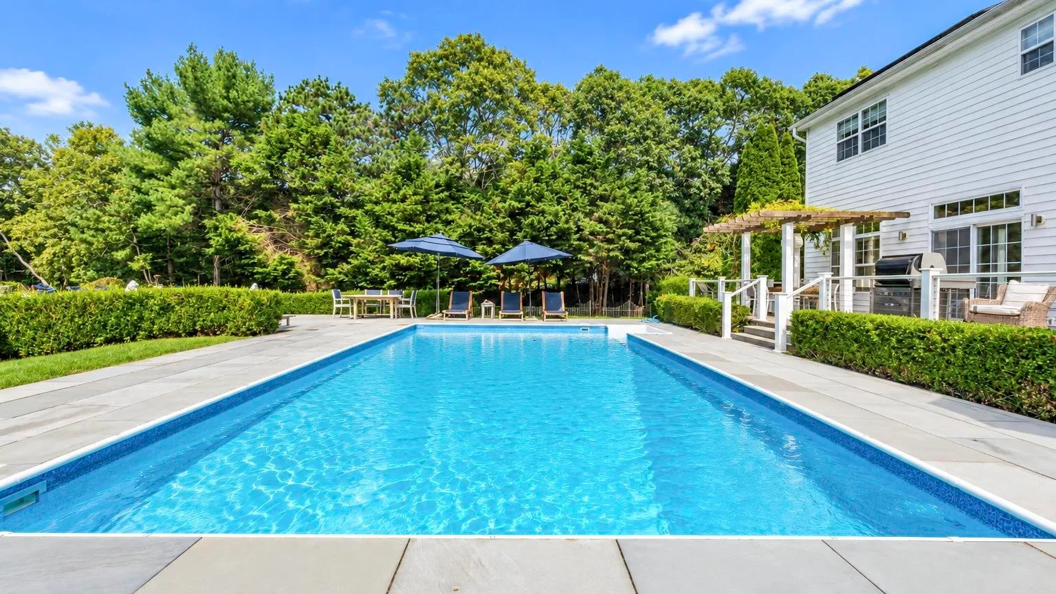 Piscina al aire libre junto a una casa blanca, con árboles al fondo.