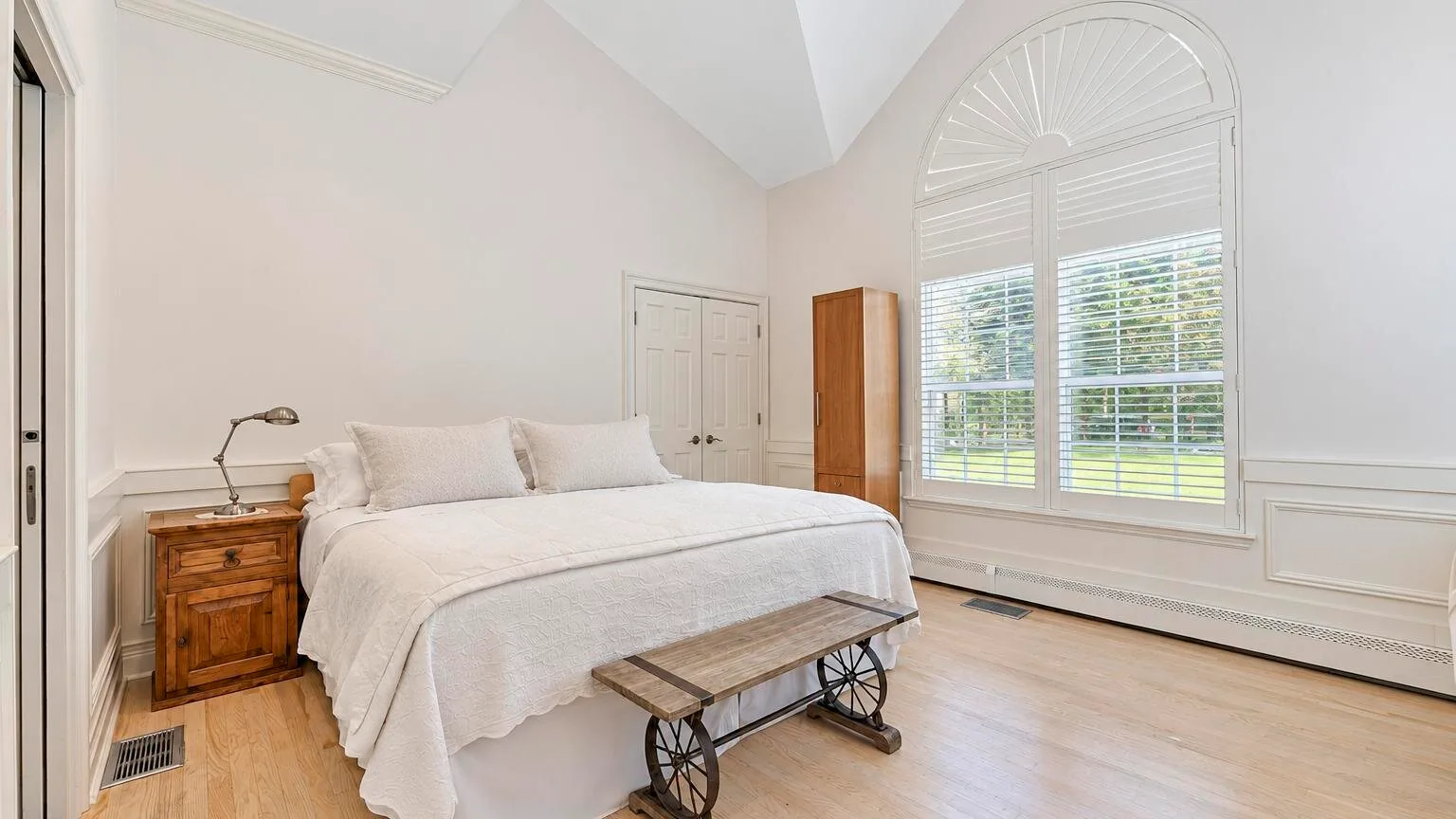 Un dormitorio luminoso con una cama blanca junto a una gran ventana arqueada.