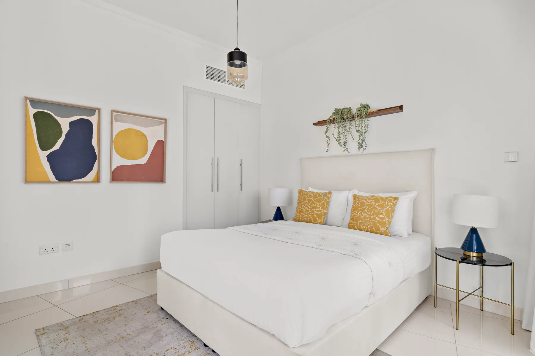 A bright bedroom with a white bed, yellow accent pillows, and colorful wall art.