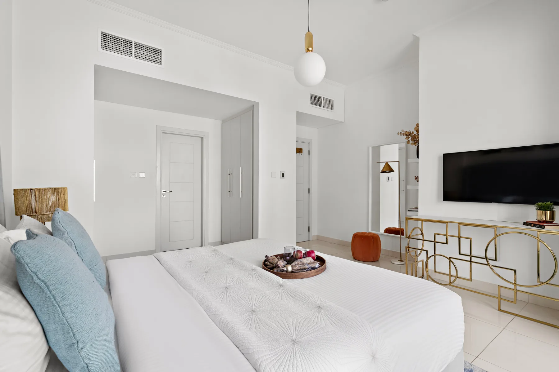 A bright bedroom with a large bed, a wall-mounted television, and a white pendant light.