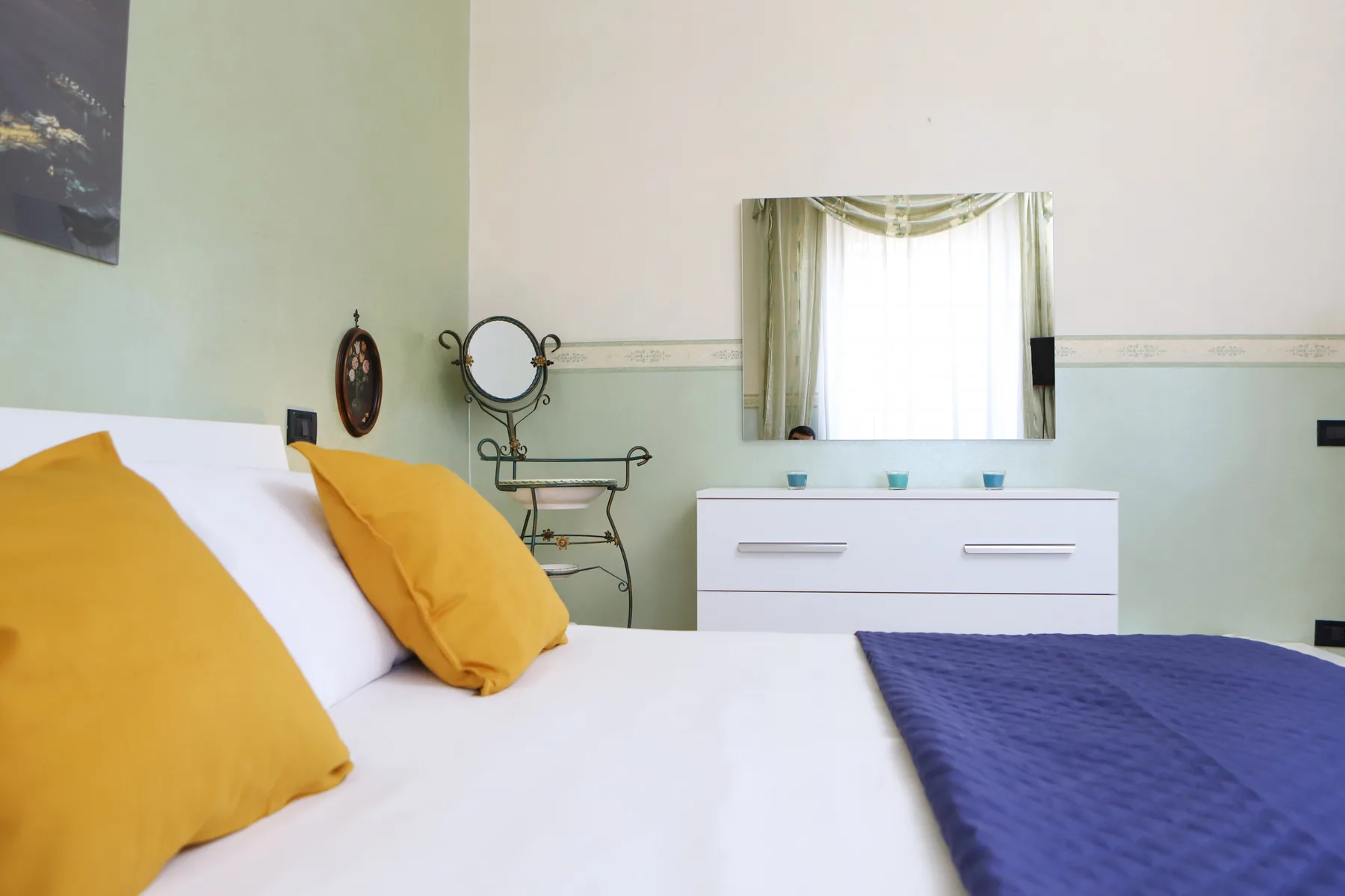 A bedroom with a white bed, colorful pillows, and a dresser beneath a window.