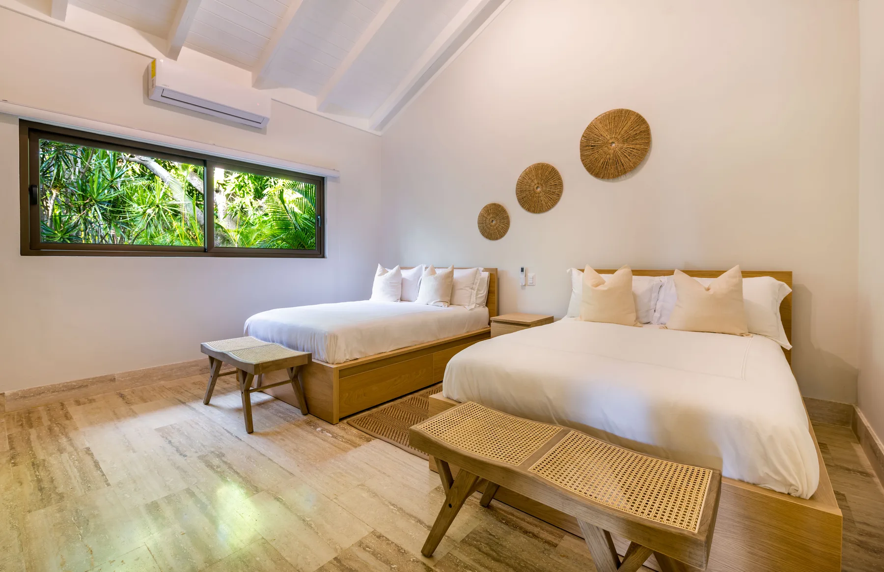 A bright bedroom with two beds and a window overlooking greenery.