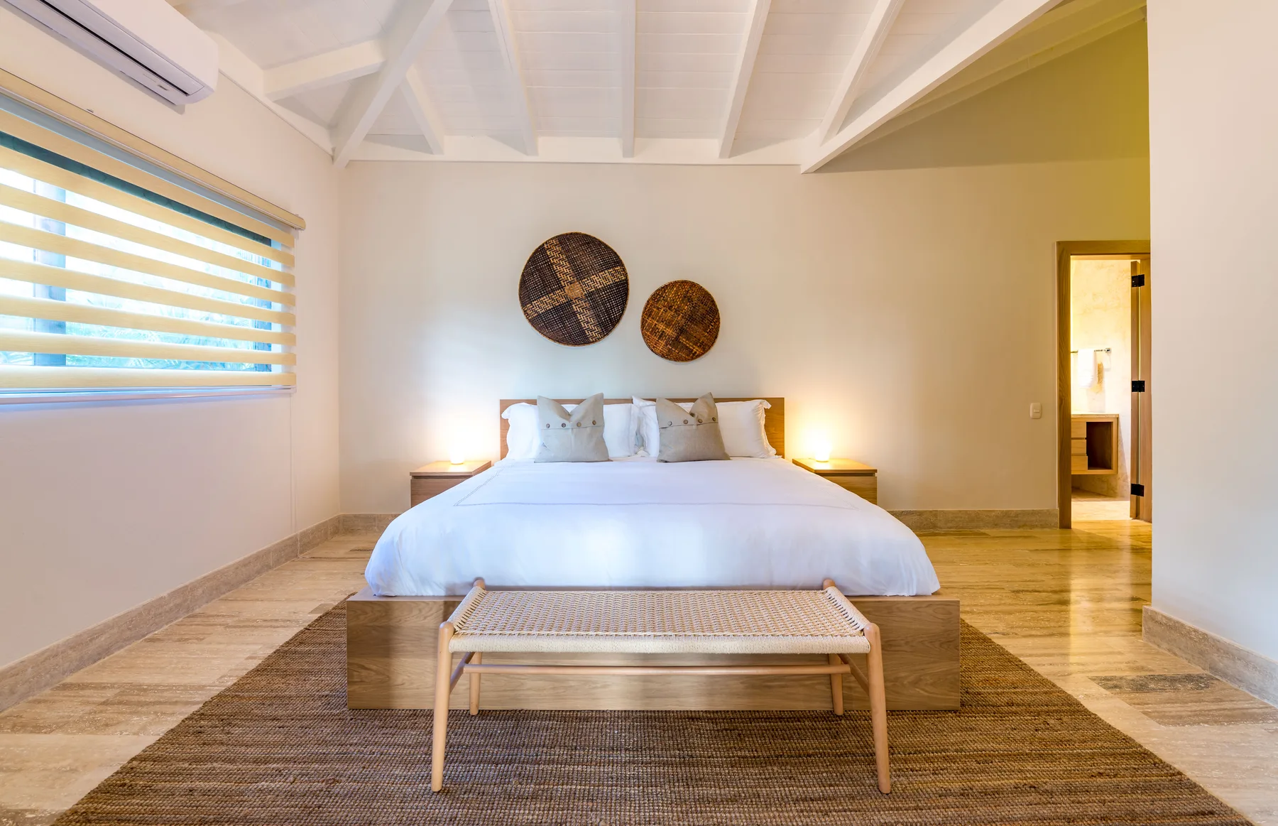 A bedroom with a white bed, woven bench, and two round wall decorations.