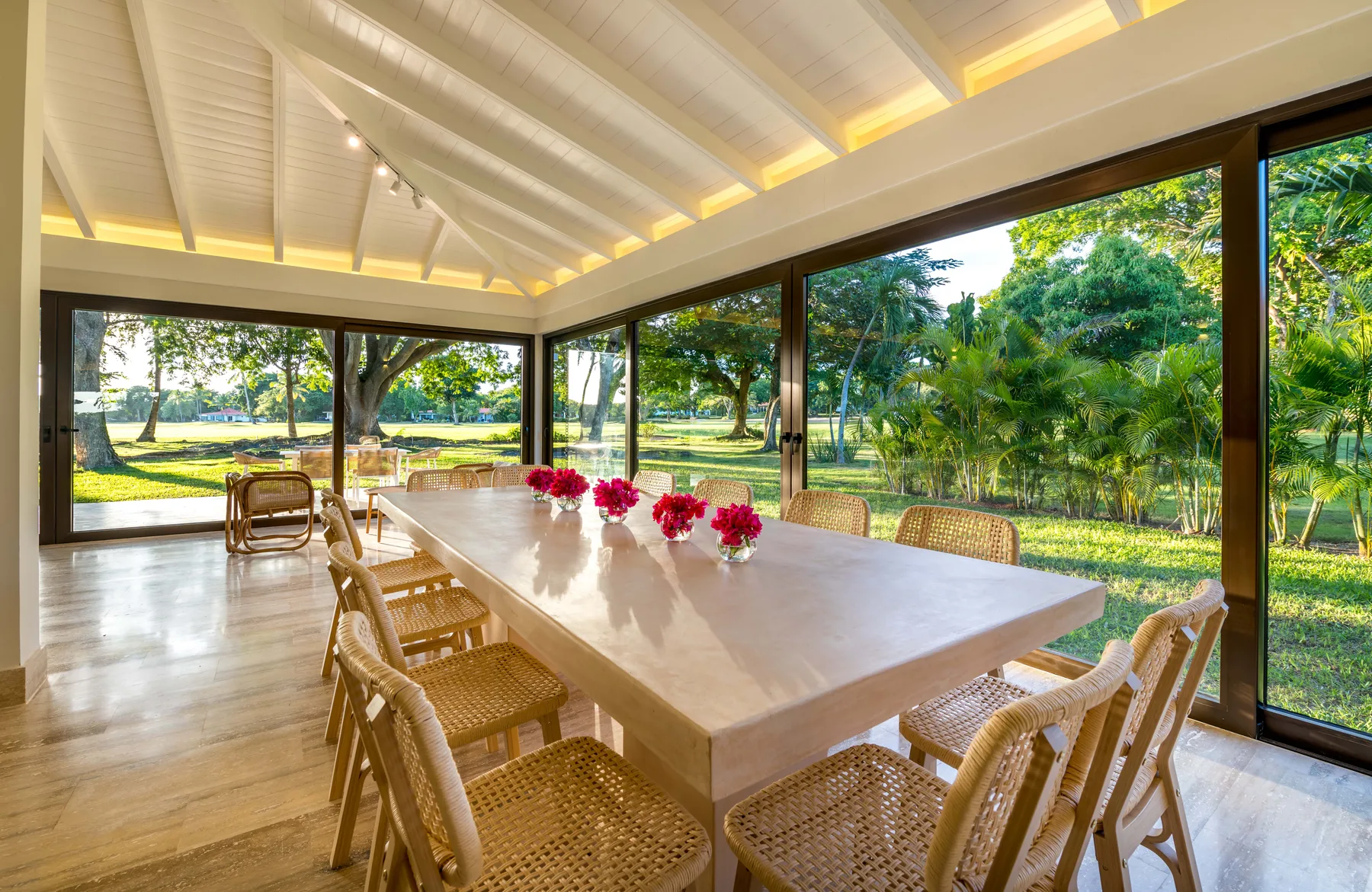 A dining area with a long table and woven chairs beside large windows.