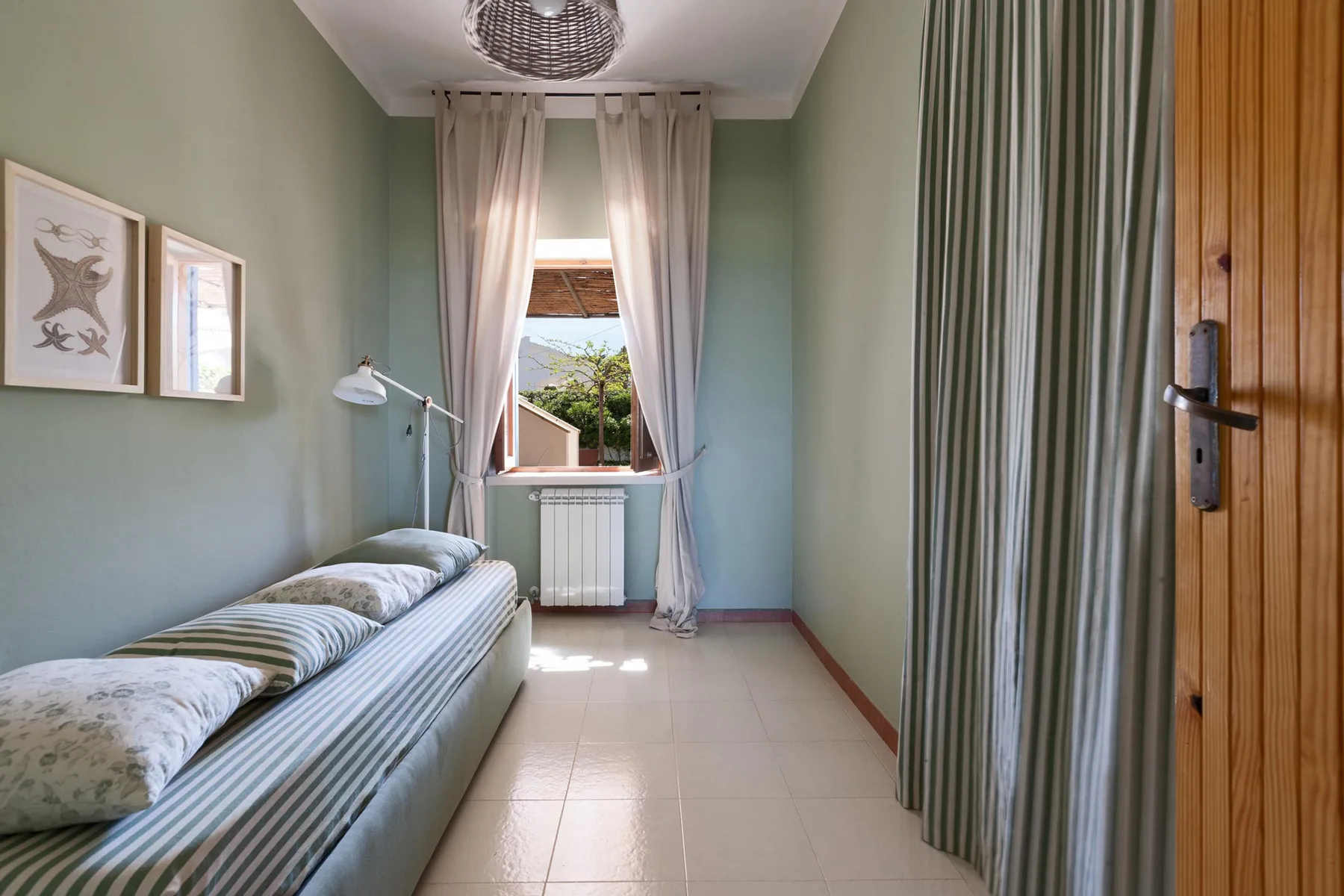 A narrow bedroom with a single bed, a curtained window, and a striped curtain.
