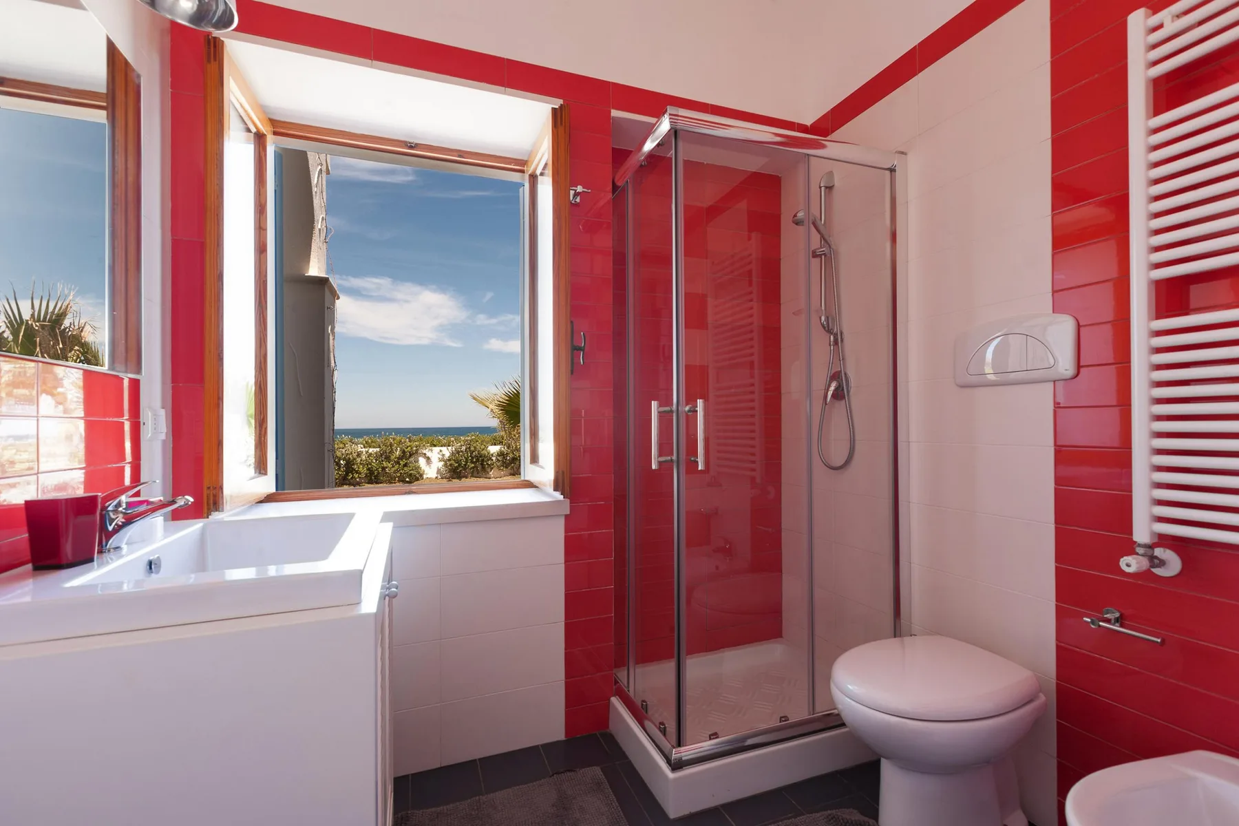 A bathroom with red tile, a glass shower, and a window overlooking the coast.