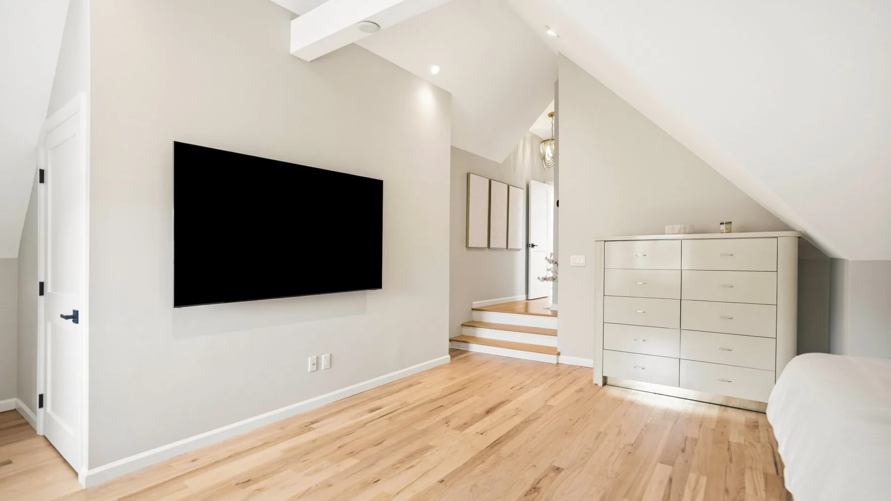 A bright attic bedroom has a wall-mounted television and a wide dresser.