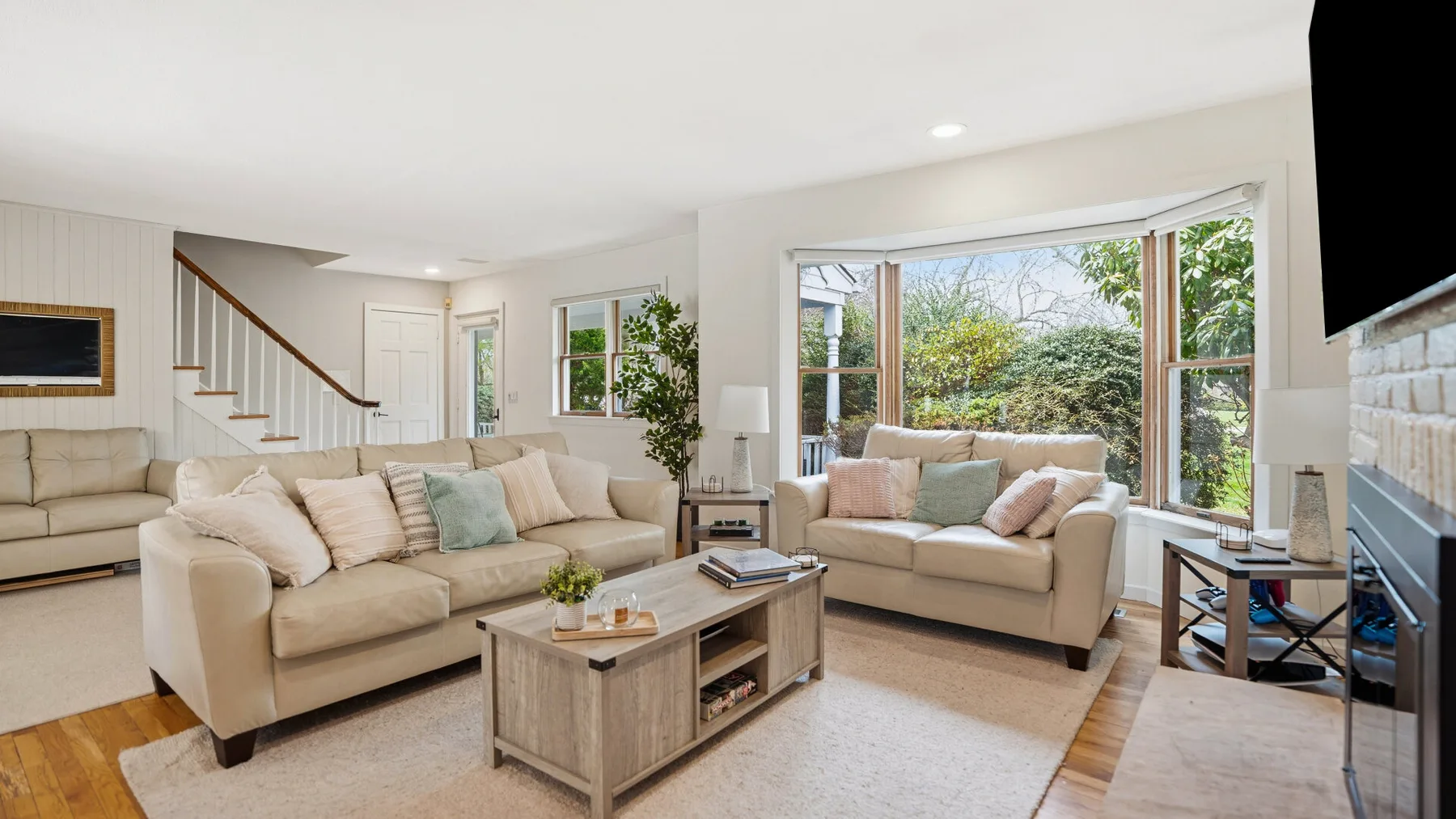 A bright living room with light-colored seating and large windows overlooking greenery.