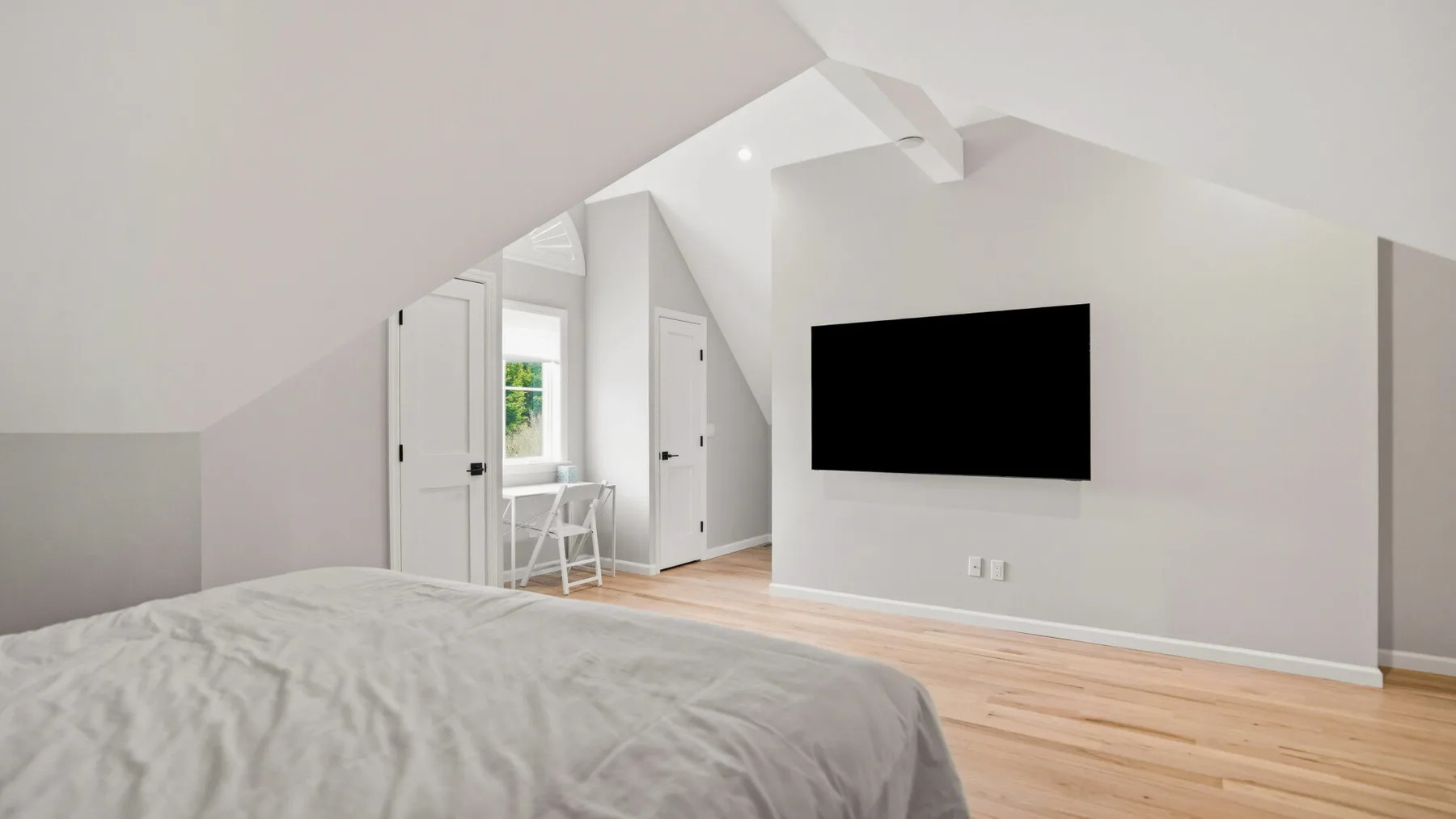 A bright bedroom with a bed, wall-mounted television, and light wood flooring.