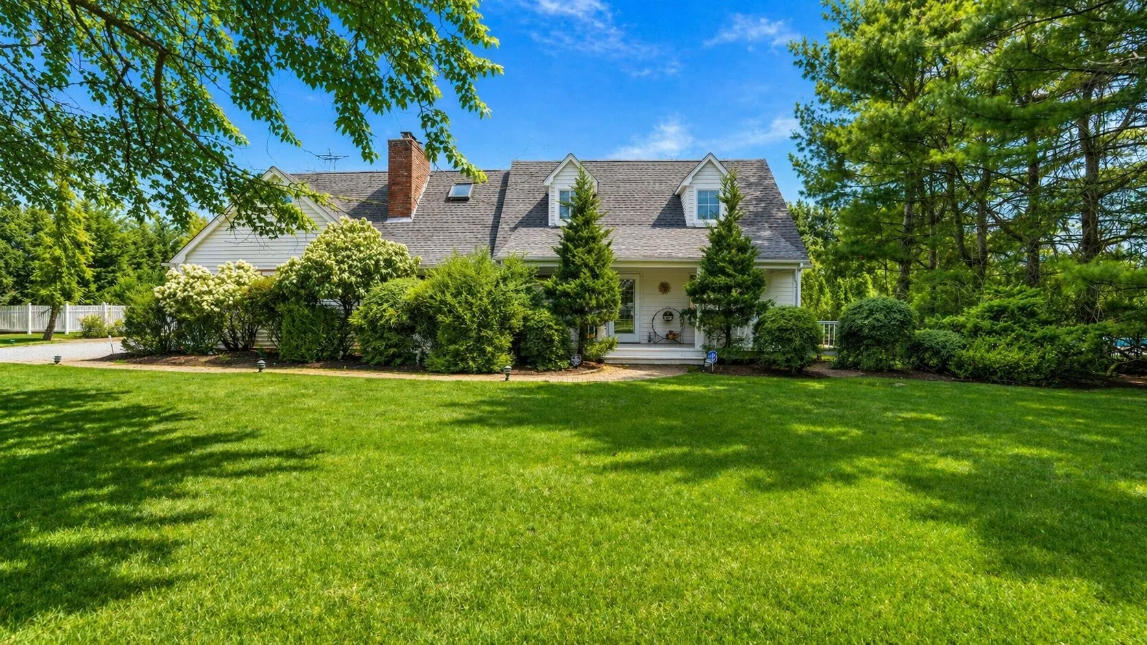 A house stands beyond a broad green lawn, surrounded by trees and shrubs.
