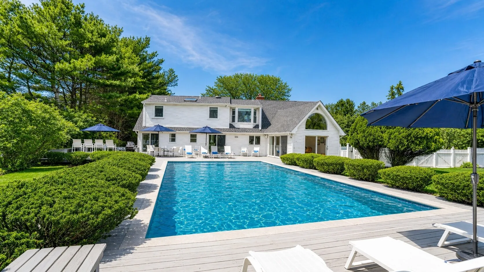 A swimming pool bordered by a wooden deck, hedges, and a large house.
