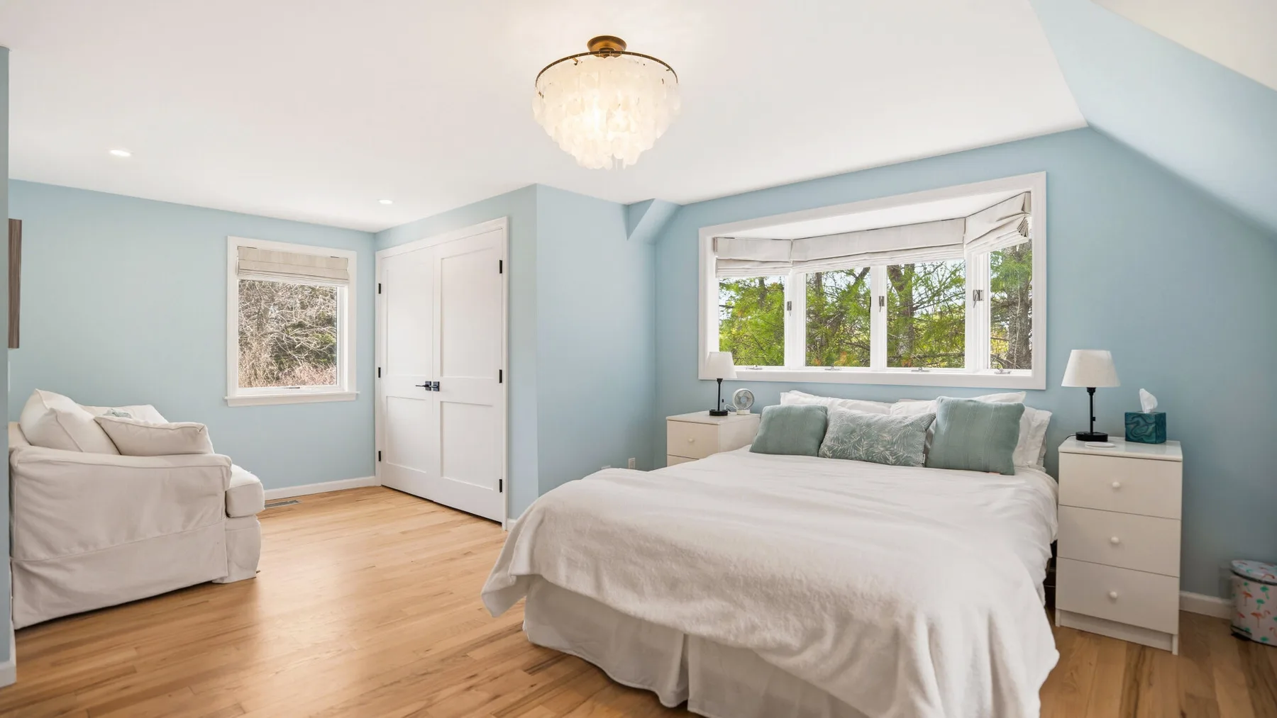 A bright bedroom with a neatly made bed, pale blue walls, and a window.