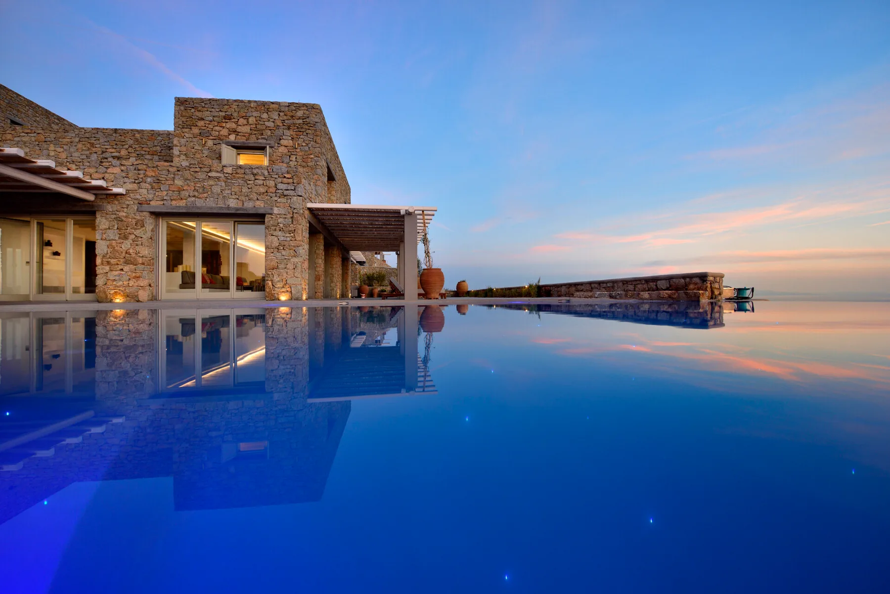 A stone villa beside an infinity pool overlooking the sea at sunset.