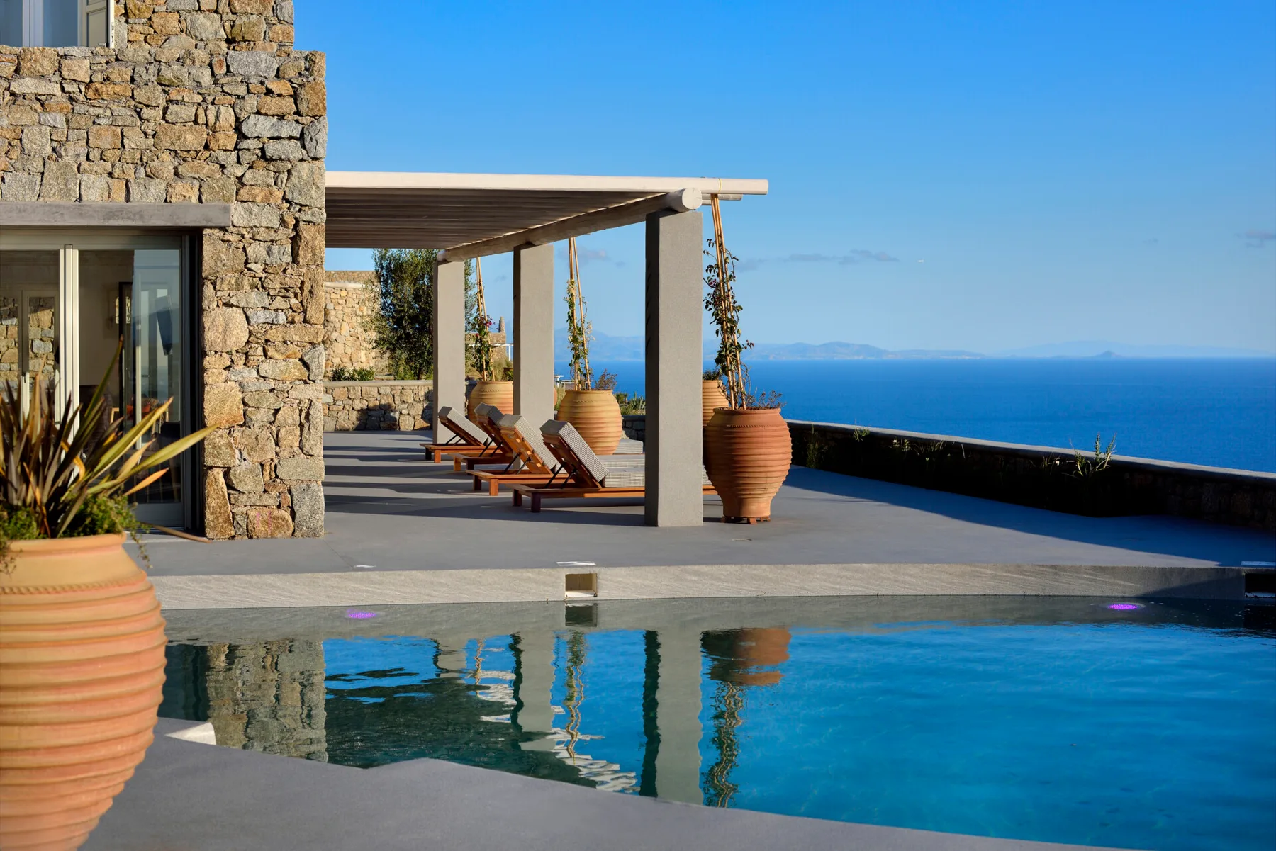 A swimming pool beside a stone villa, with a shaded seating area overlooking the sea.