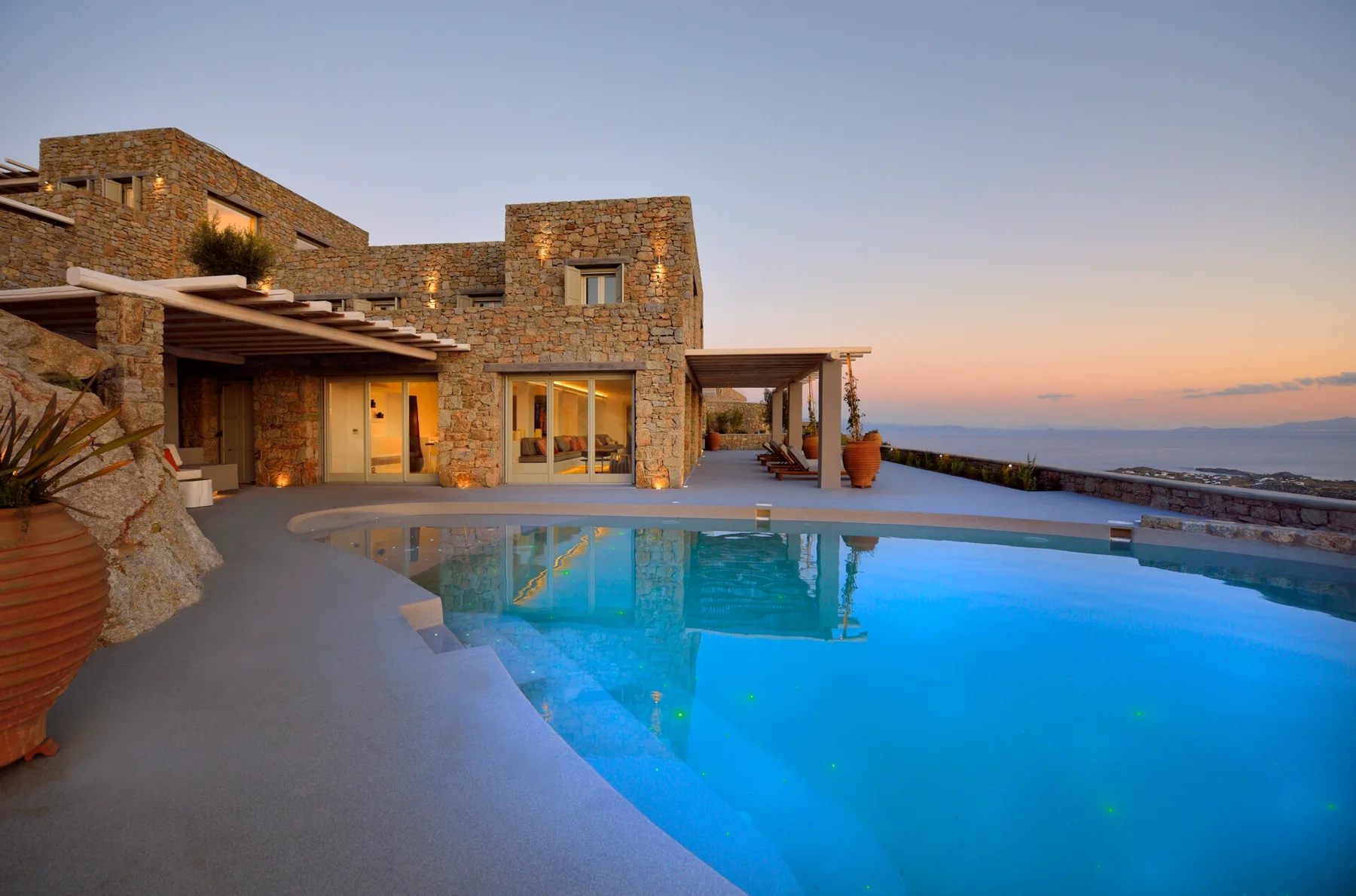 Stone villa beside a swimming pool overlooking the sea at sunset.