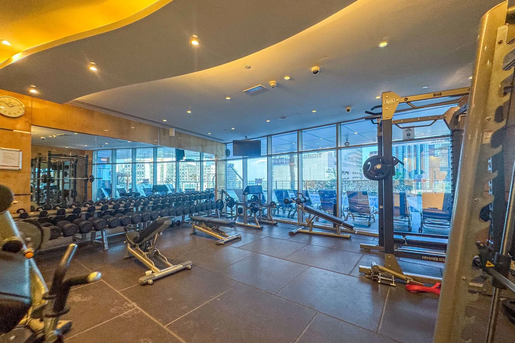 A gym with exercise machines and dumbbells beside large windows.