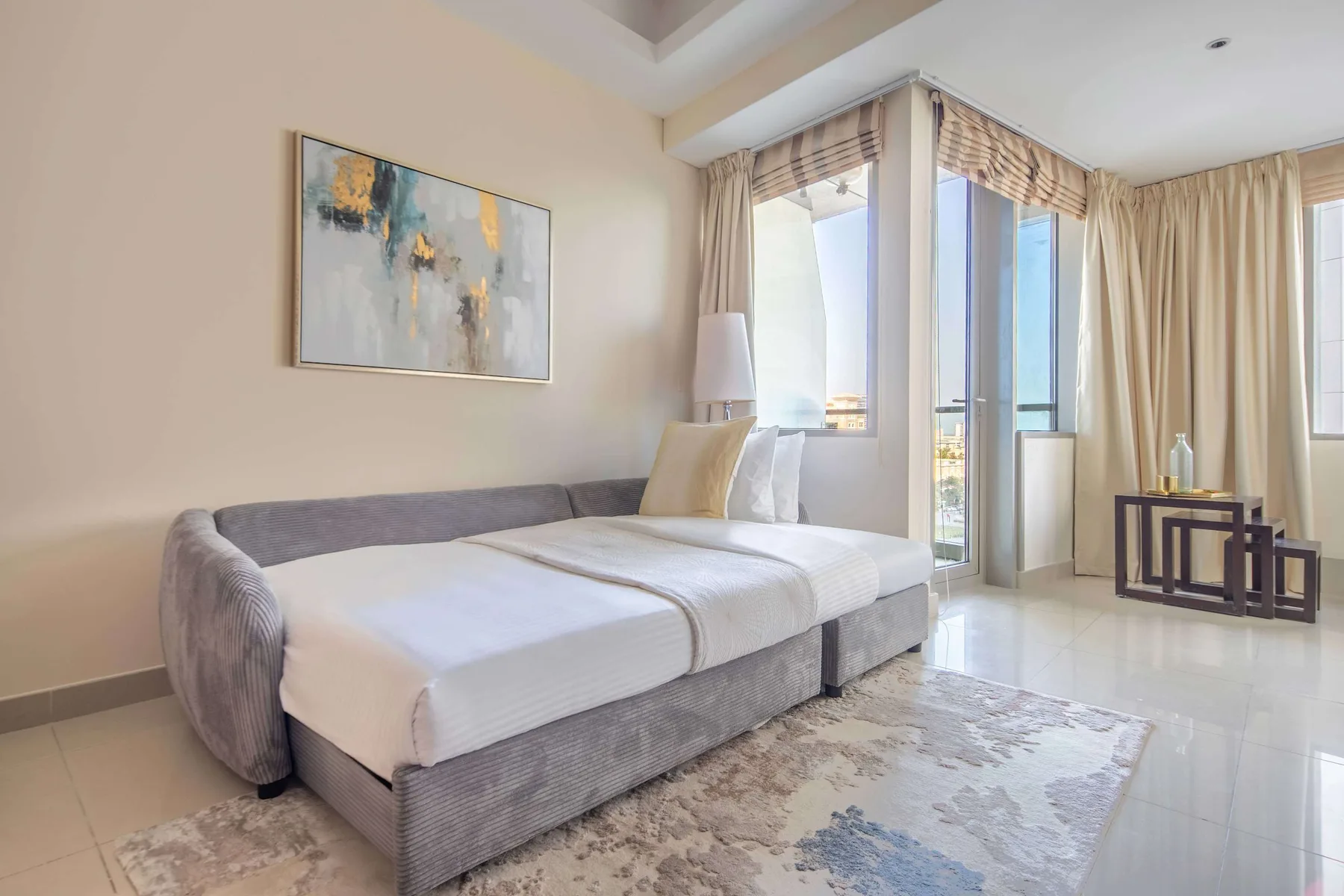 A bedroom with a neatly made bed beside tall windows and light curtains.