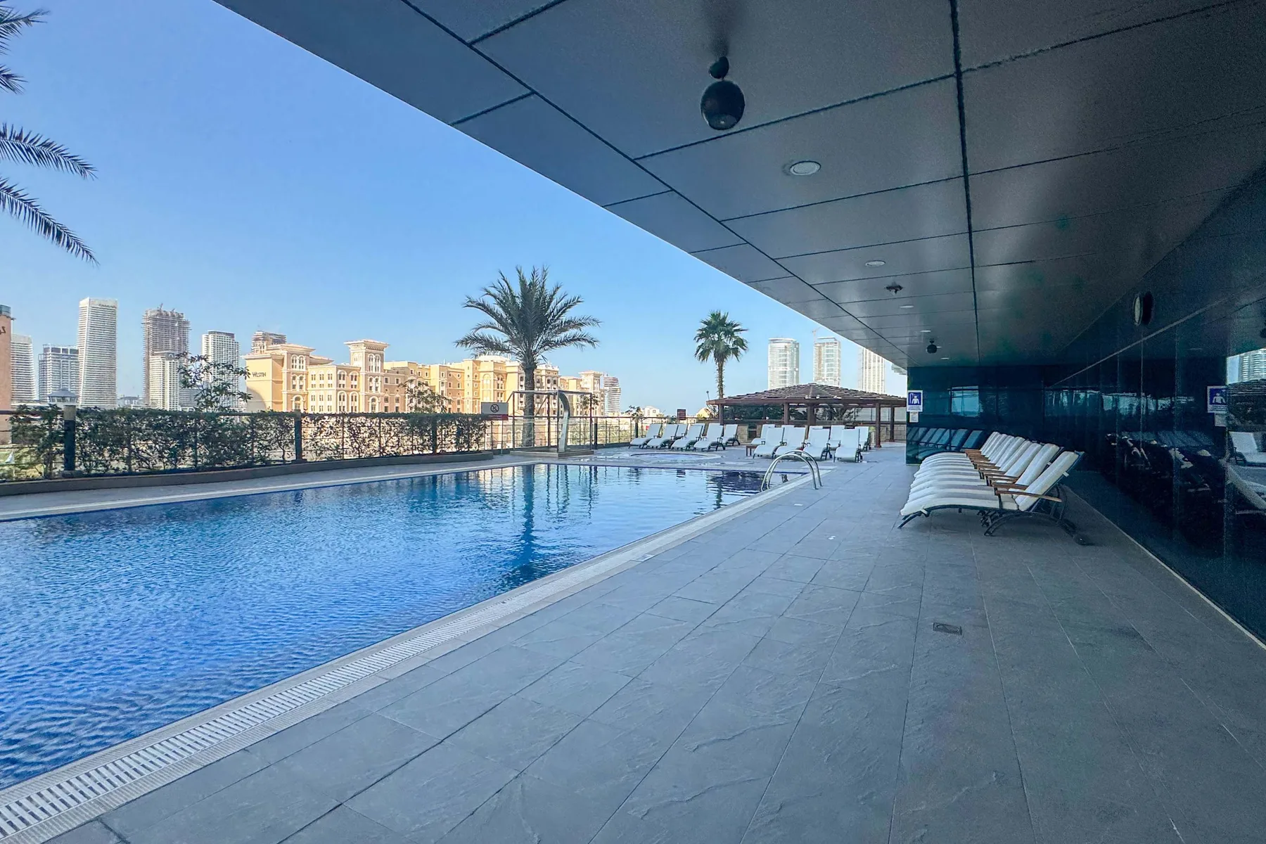 A rooftop swimming pool sits beside a shaded deck with lounge seating and city buildings beyond.