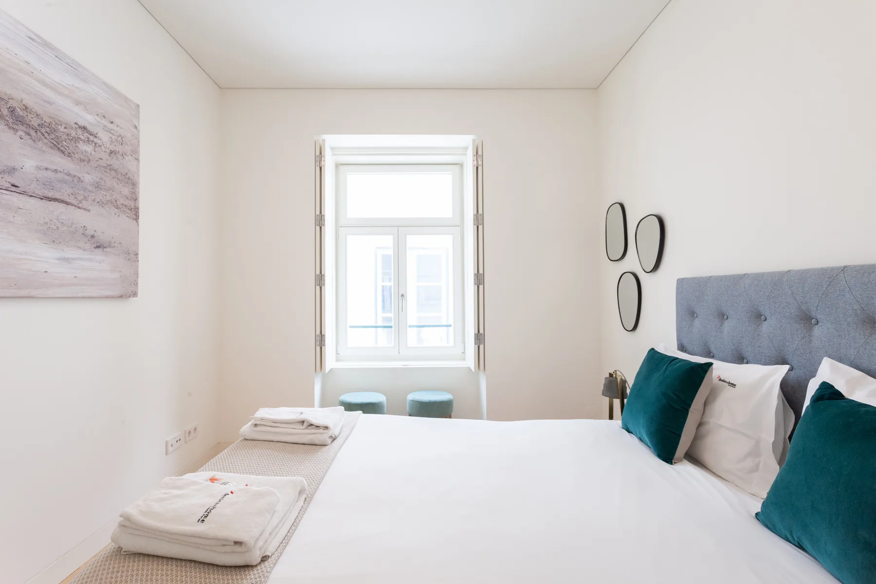 Dormitorio con una cama, cojines turquesa y una ventana luminosa.