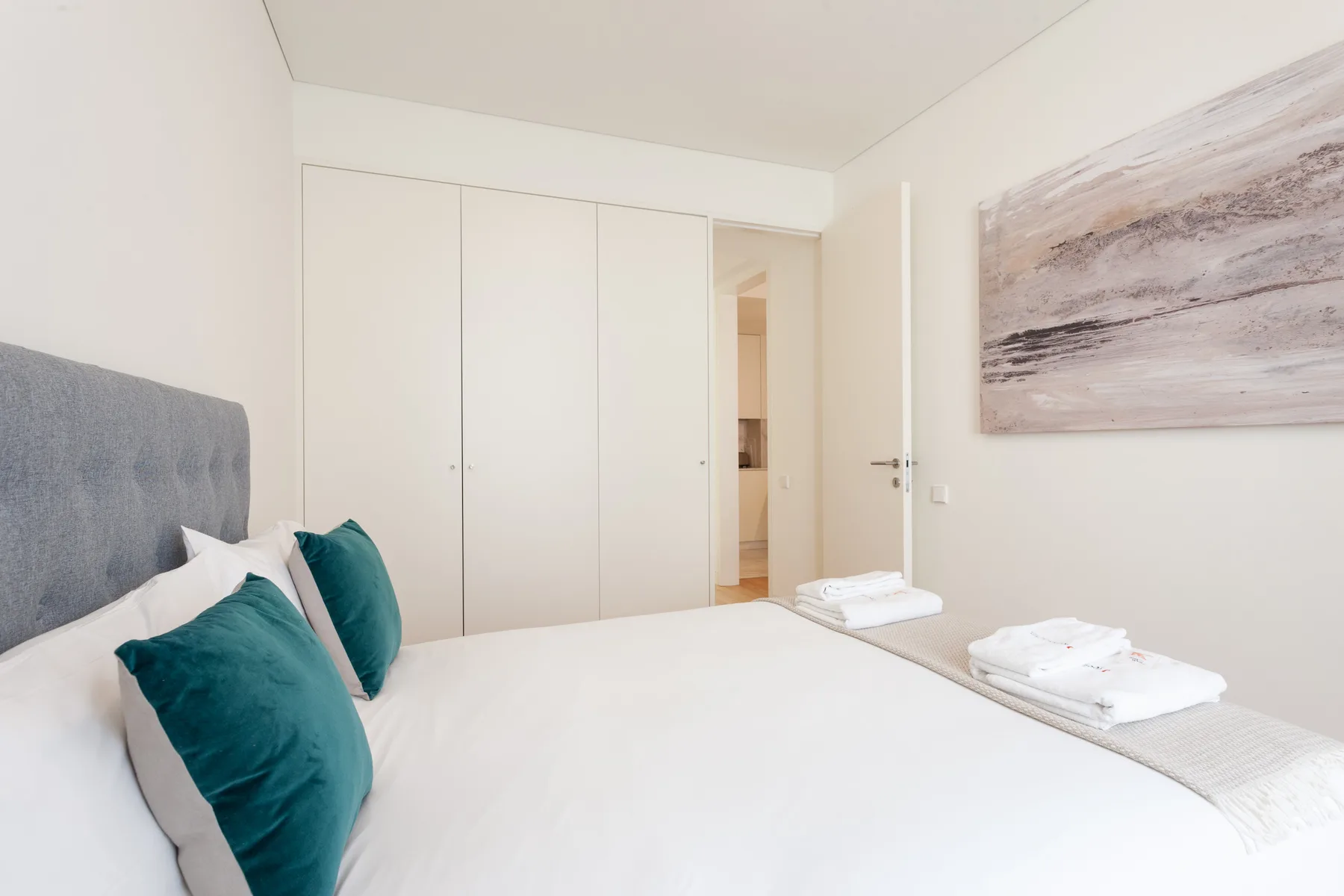 Un dormitorio con una cama blanca, cojines verde azulado y un cuadro de paisaje en la pared.