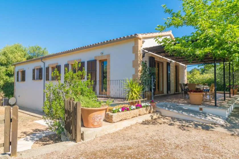 Una casa blanca con una zona de estar al aire libre a la sombra y parterres con plantas.