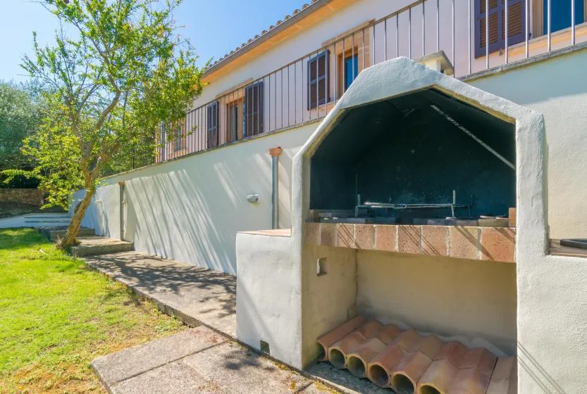 Una zona de barbacoa al aire libre junto a una casa blanca, con un árbol frondoso cerca.