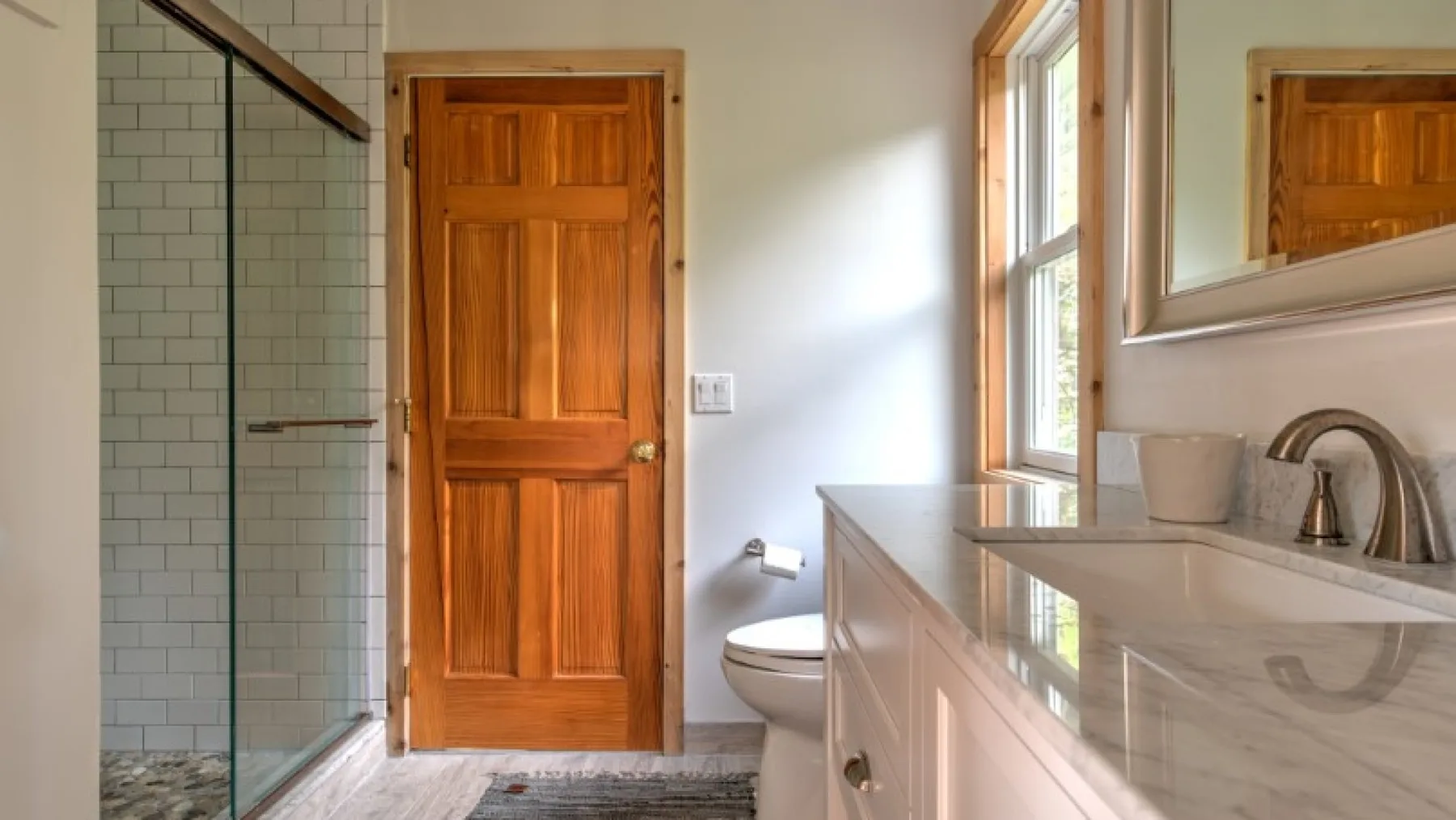 Un baño con ducha de vidrio, puerta de madera, inodoro y tocador con lavabo.