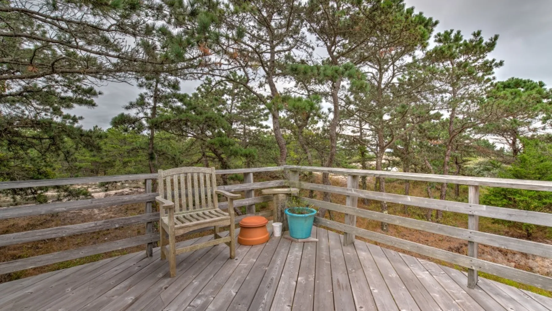 Una terraza de madera con barandillas da a los árboles, con macetas junto a un banco.