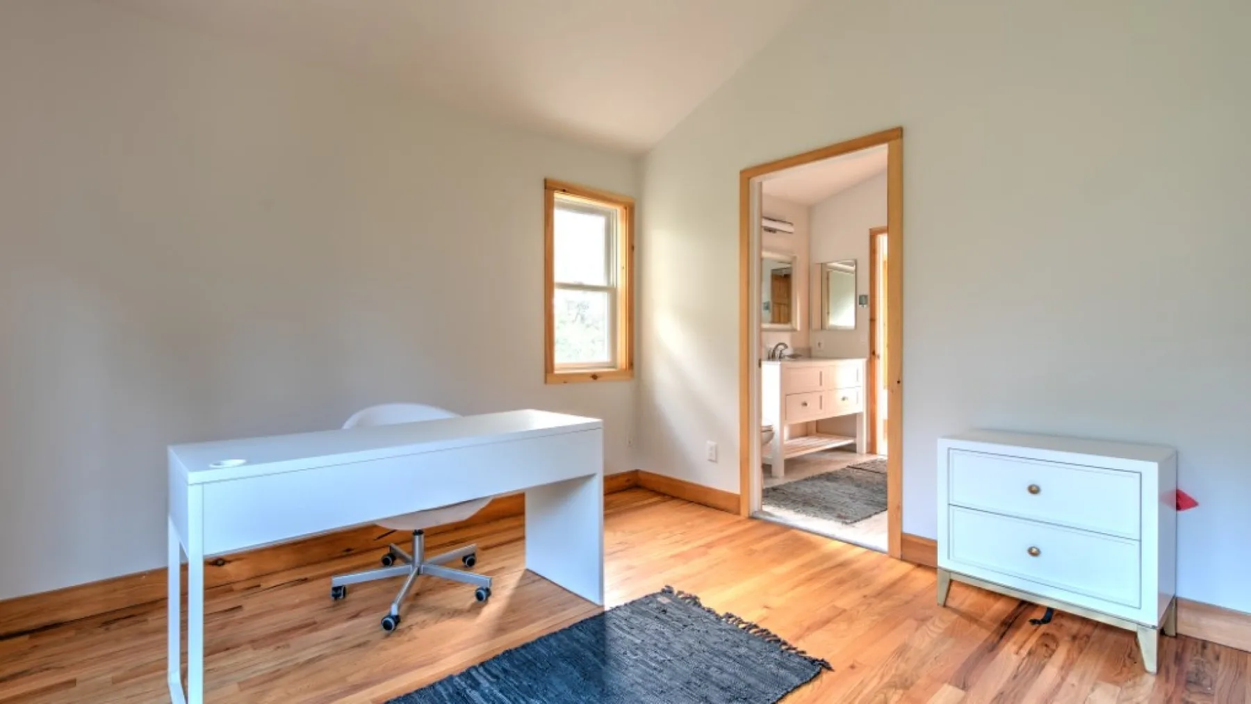 Una habitación luminosa con un escritorio blanco, un mueble pequeño y una puerta a otra estancia.
